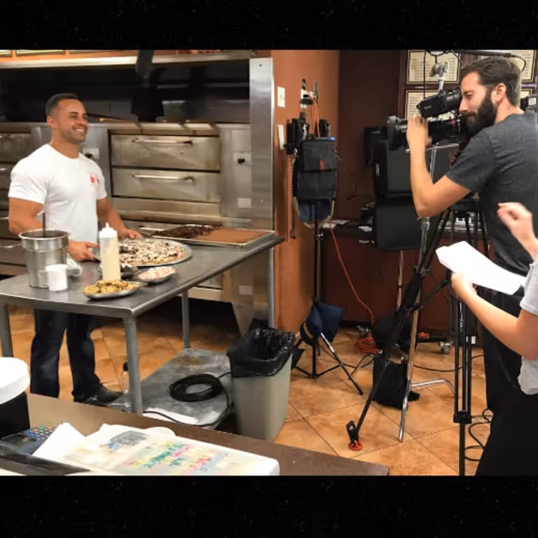 A man smiles in a pizzeria kitchen, presenting pizzas on a table. A cameraman films him nearby.