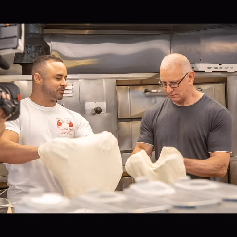 Two men in a kitchen stretch pizza dough. One wears a white shirt with a pizzeria logo, the other a gray shirt. They focus intently on their work.