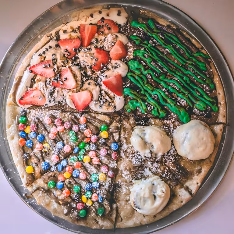 Dessert pizza divided into four sections topped with strawberries and cream, green icing, colorful candy-coated chocolates, and meringue cookies with powdered sugar.