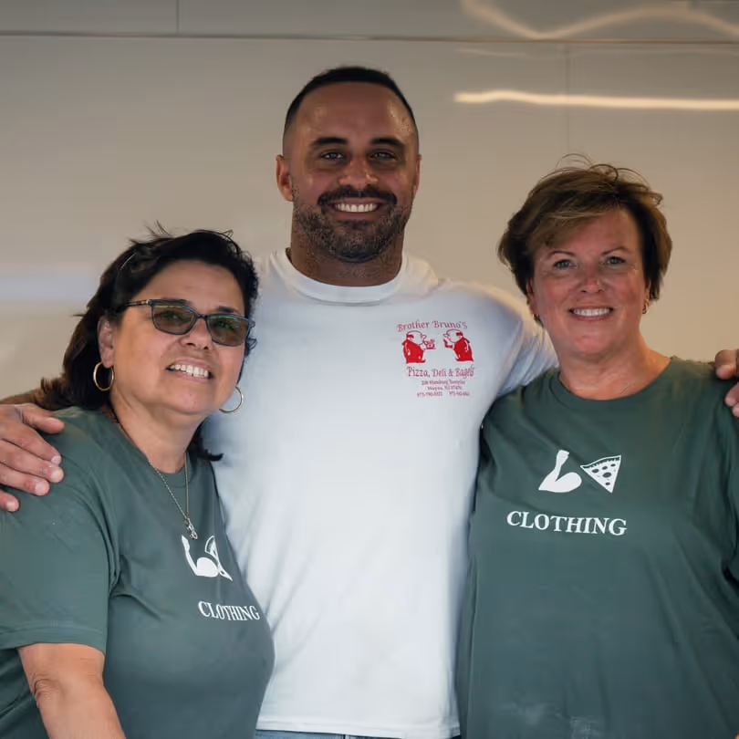 Three people smiling and posing together, with two women wearing matching green shirts with a flexed arm and pizza slice design and the word 'CLOTHING', and a man in the center wearing a white shirt with Brother Bruno's Pizza Deli & Bagels logo.