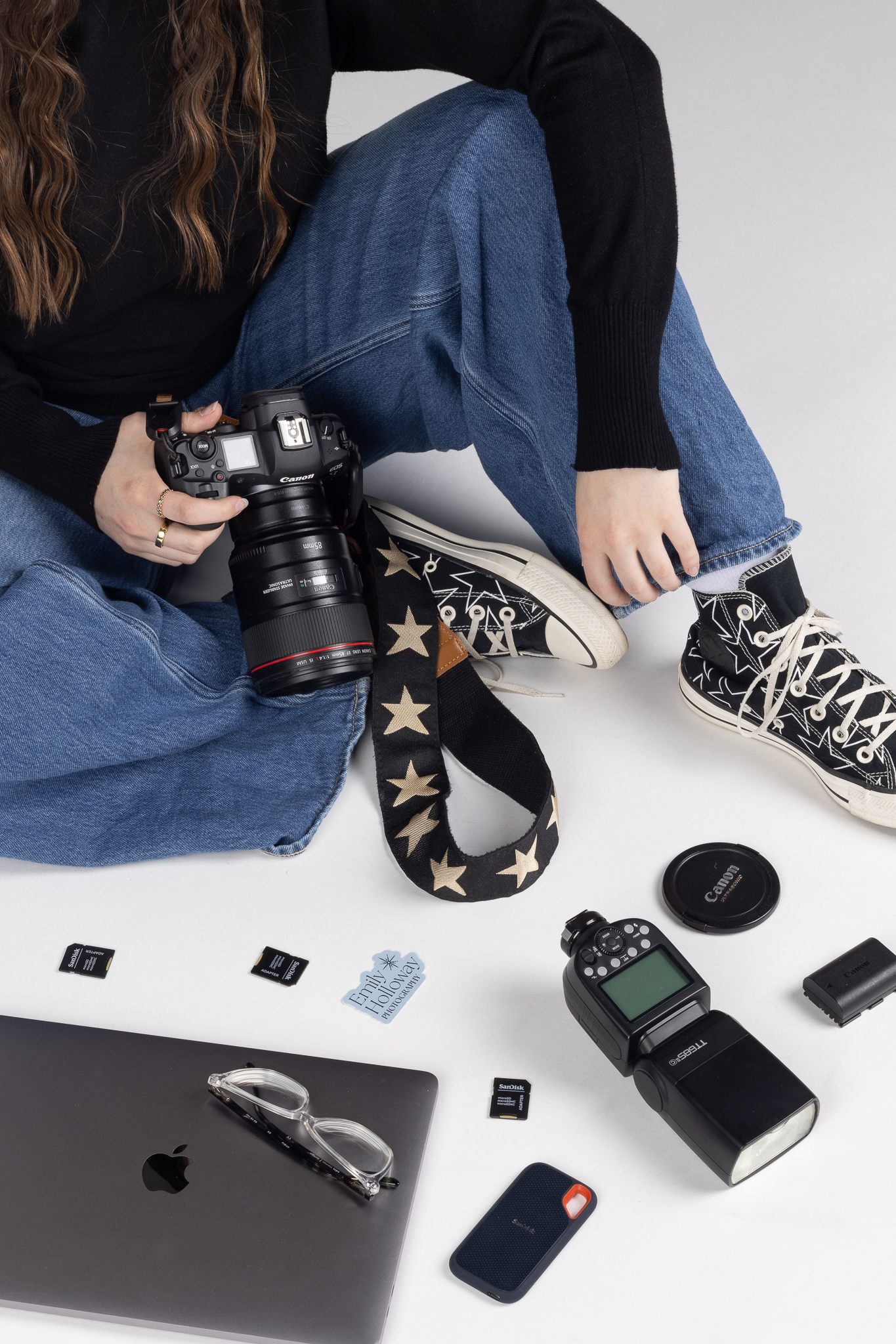 Person sitting cross-legged holding a Canon camera with a star-patterned strap, surrounded by photography accessories including memory cards, a flash, lens cap, battery, a closed MacBook with glasses on top, and an external SSD.