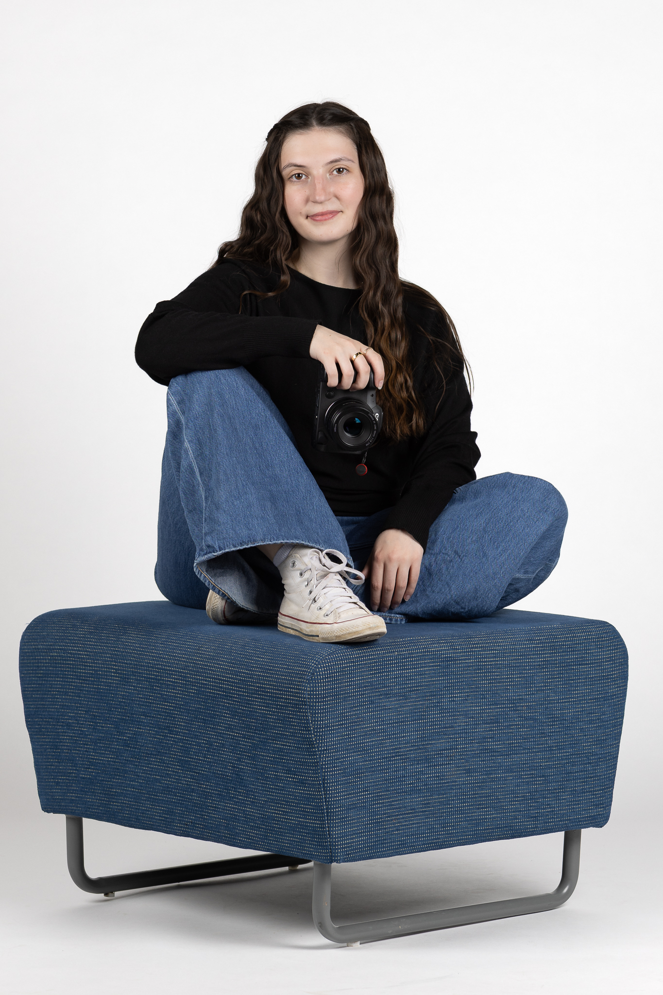 Young woman with long wavy hair sitting cross-legged on a blue ottoman, holding a camera and wearing a black sweater, blue jeans, and white sneakers.
