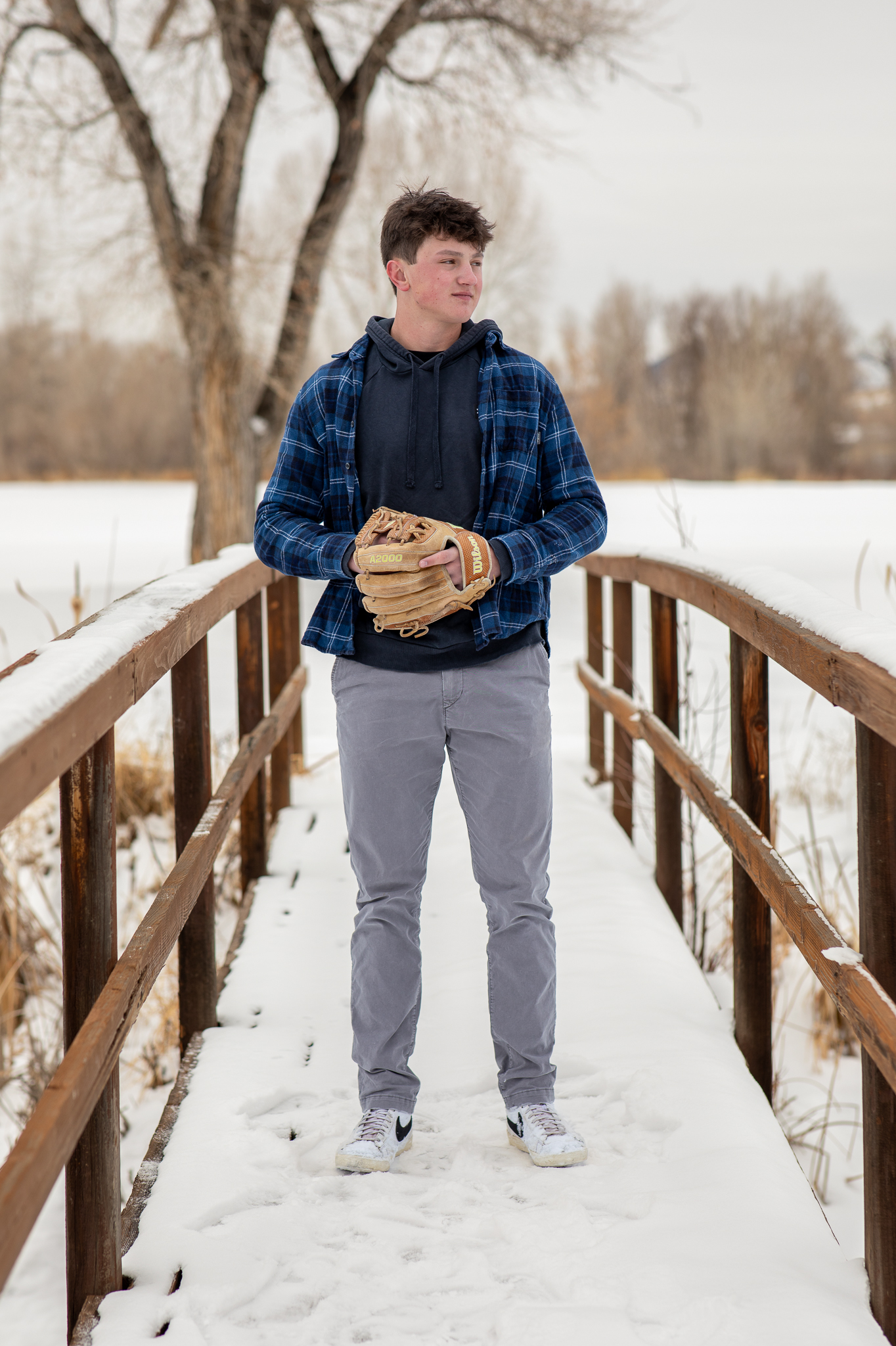 Teenage boy standing on a snow-covered wooden bridge holding a baseball glove, wearing a blue plaid shirt and gray pants.
