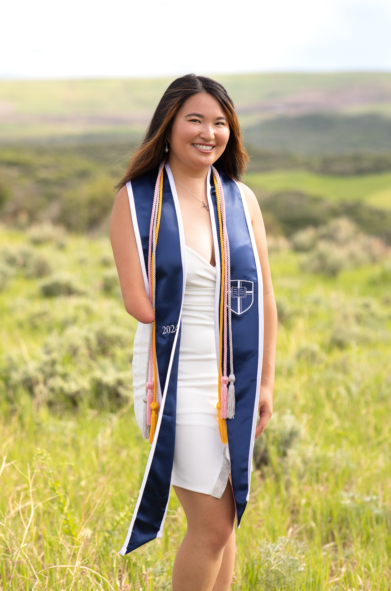 Smiling young woman in a white dress with a blue graduation stole and honor cords standing in a green field.