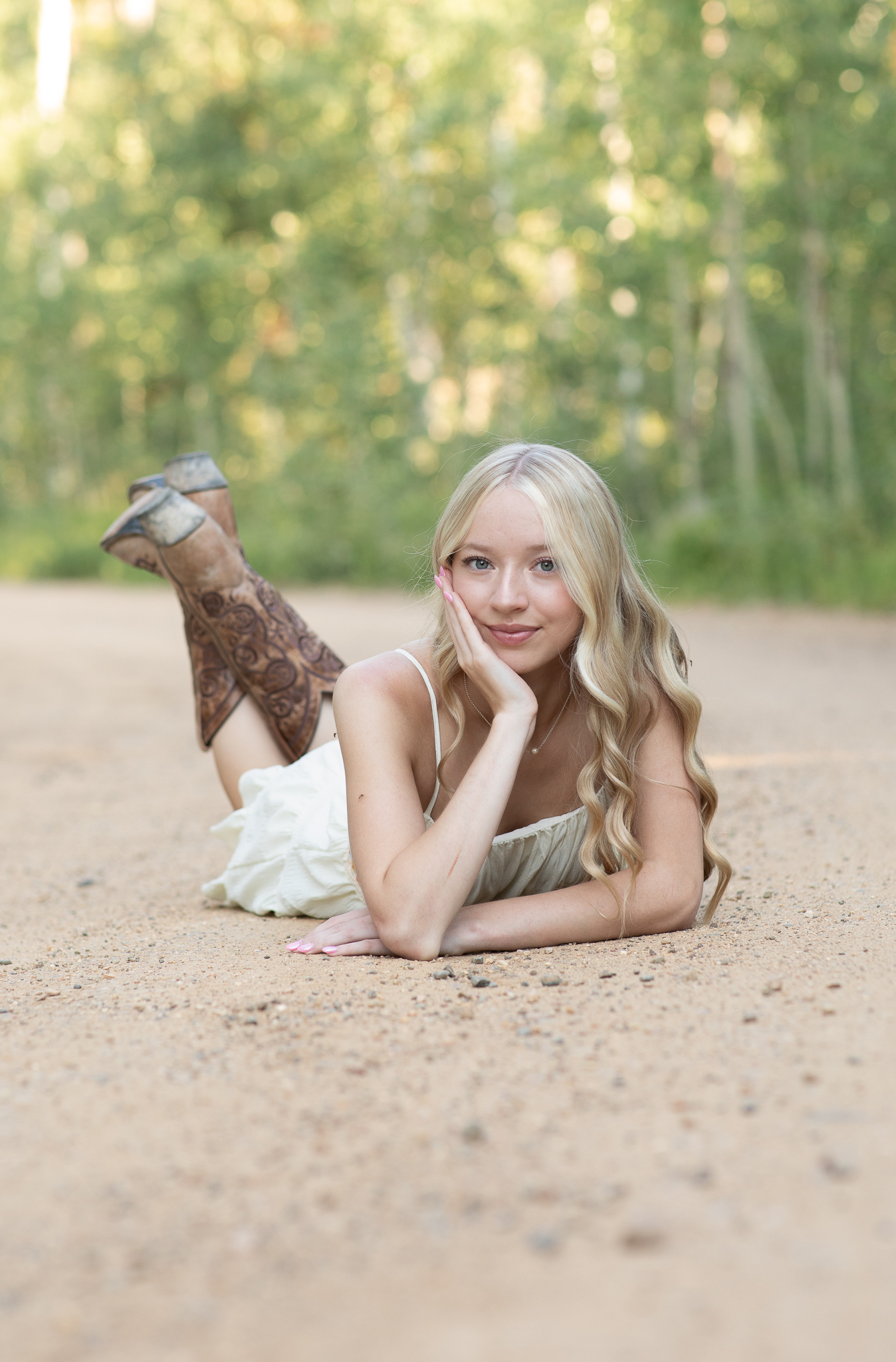 Young woman with long blonde hair lying on her stomach on a dirt road wearing a white dress and brown cowboy boots, resting her face on one hand.