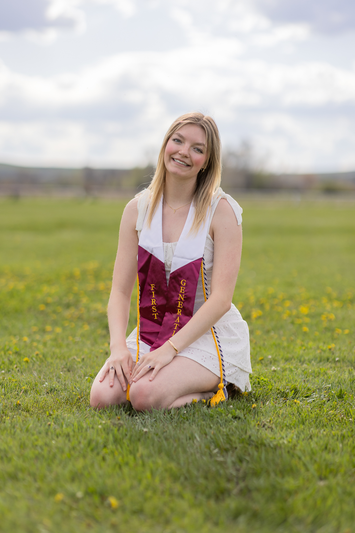 Smiling young woman kneeling on grass wearing a white dress and a maroon and white graduation stole that reads 'First Generation'.