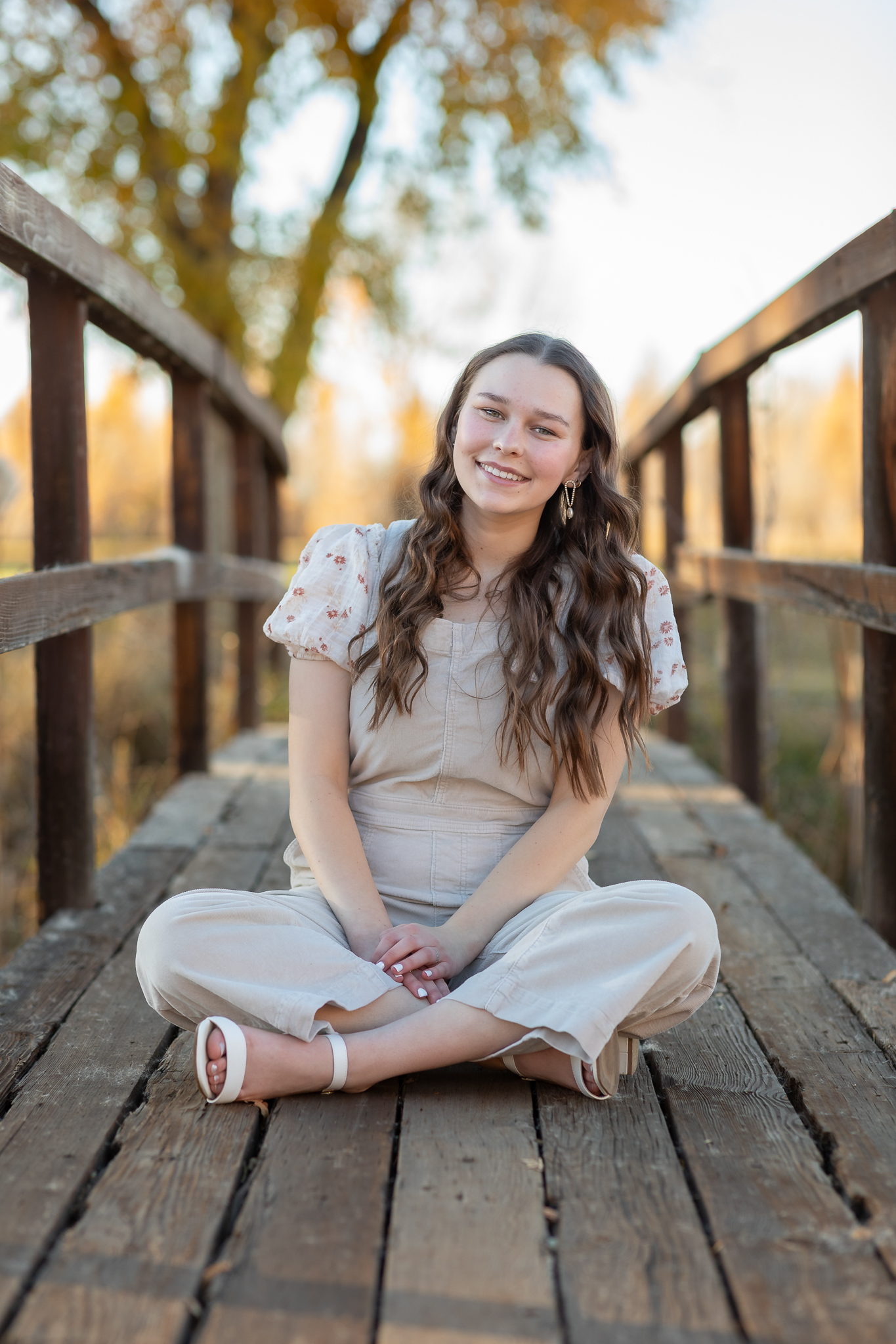 Smiling young woman with long wavy hair sitting cross-legged on a wooden bridge in a sunlit outdoor setting.