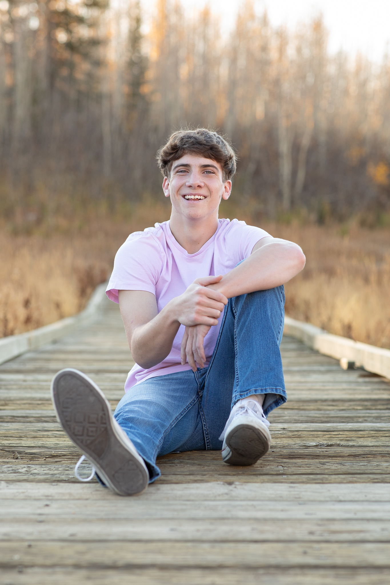 Smiling teenage boy in a light purple shirt and jeans sitting on a wooden boardwalk with trees in the background.