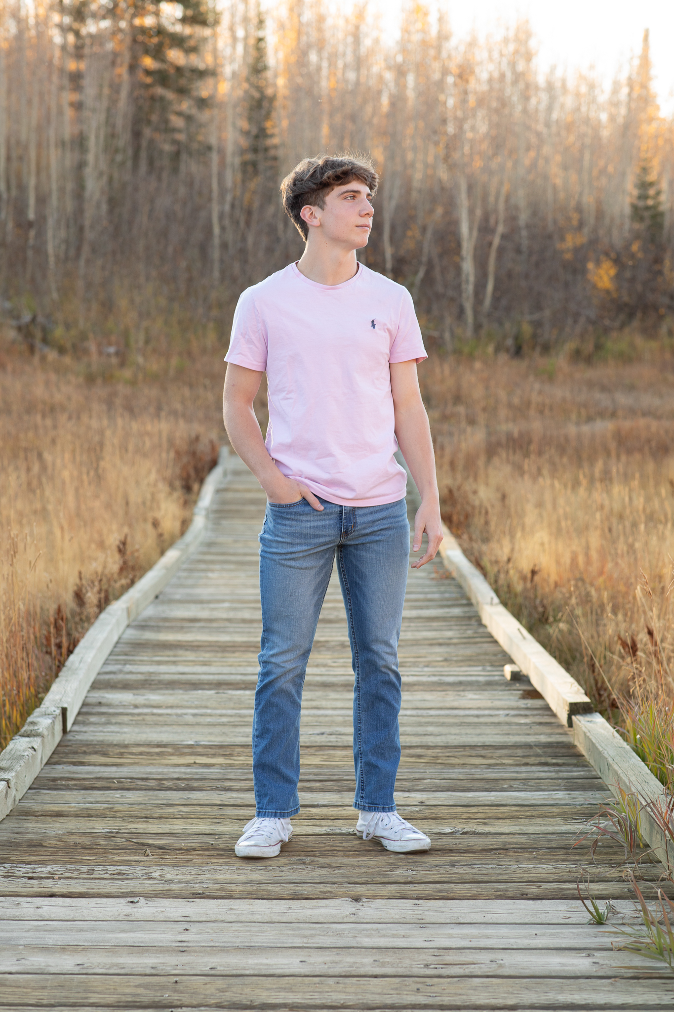 Teenage boy in a pink T-shirt and blue jeans standing on a wooden boardwalk in a field with autumn trees in the background.