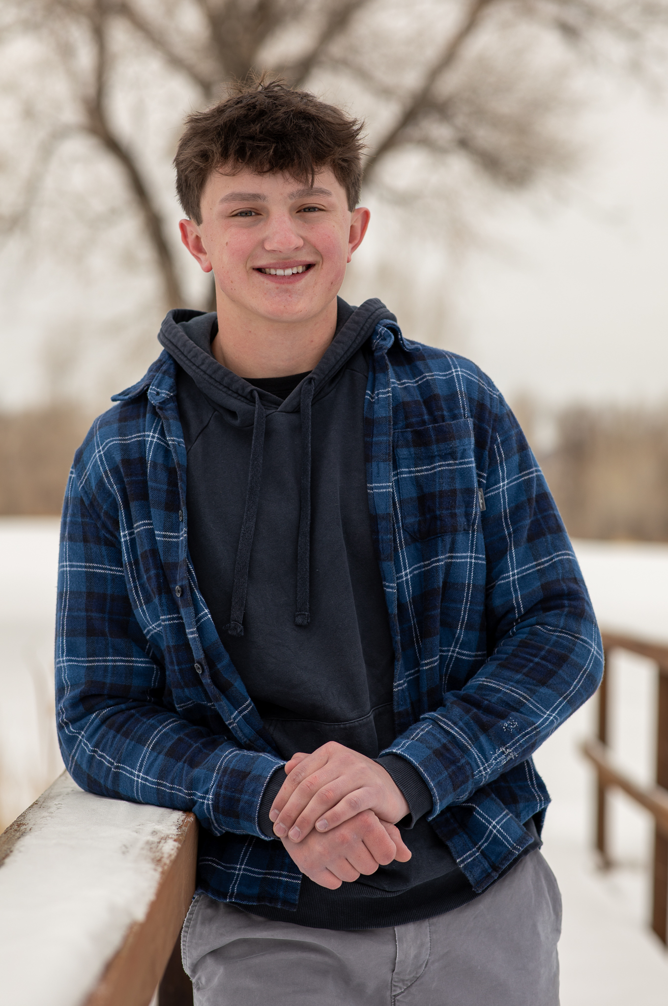 Smiling young man wearing a blue plaid shirt and black hoodie leaning on a snowy wooden railing outdoors in winter.
