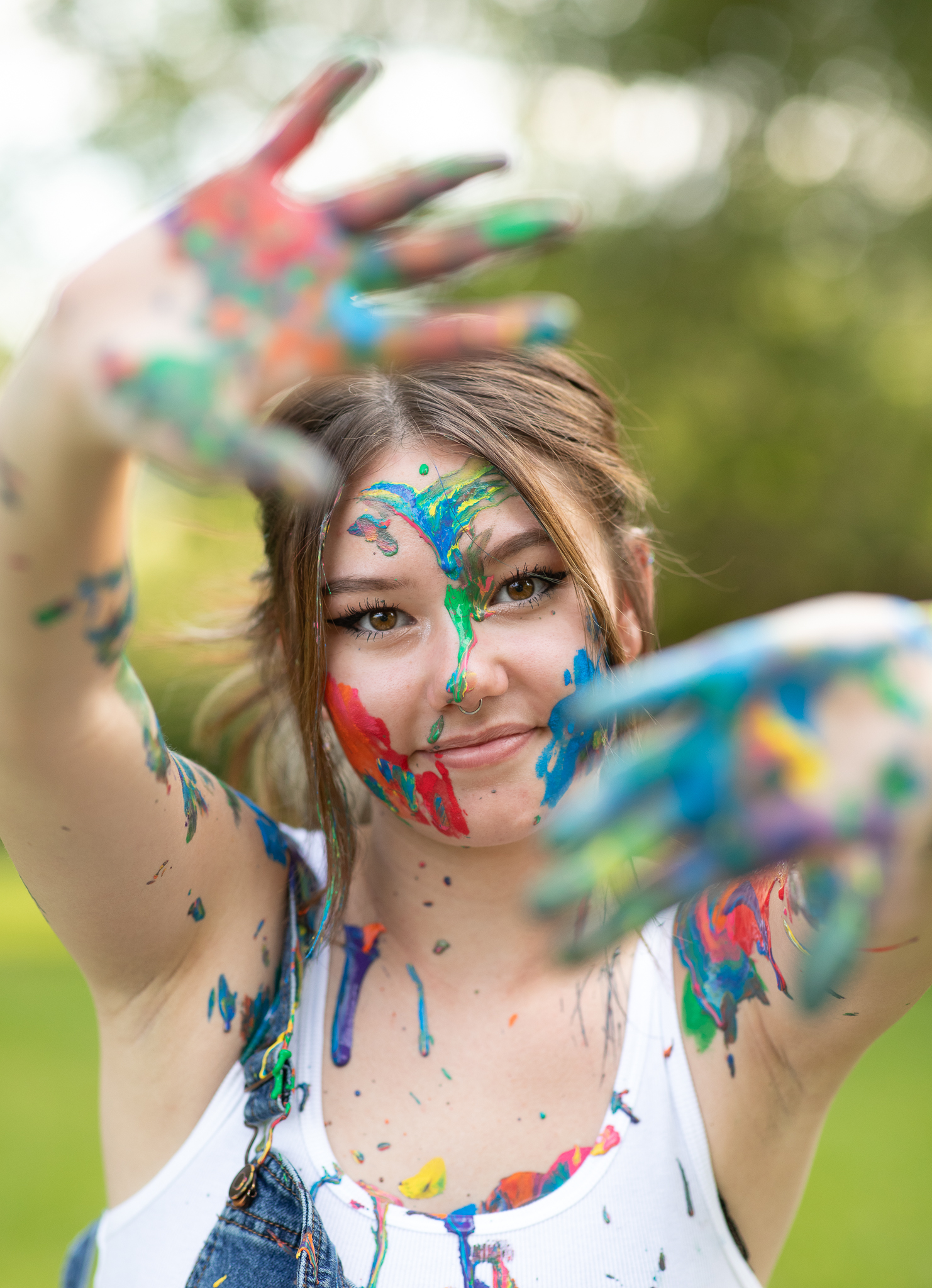 Young woman with paint splattered on her face, hands, and white tank top smiling outdoors with blurred greenery in background.