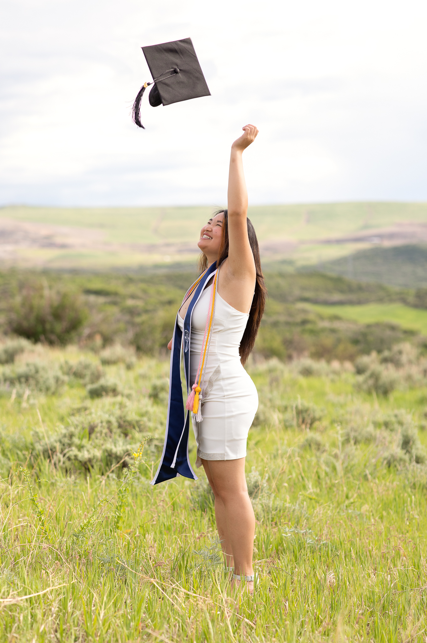 Smiling female graduate wearing a white dress and graduation stole tossing her cap outdoors in a grassy field with hills in the background.
