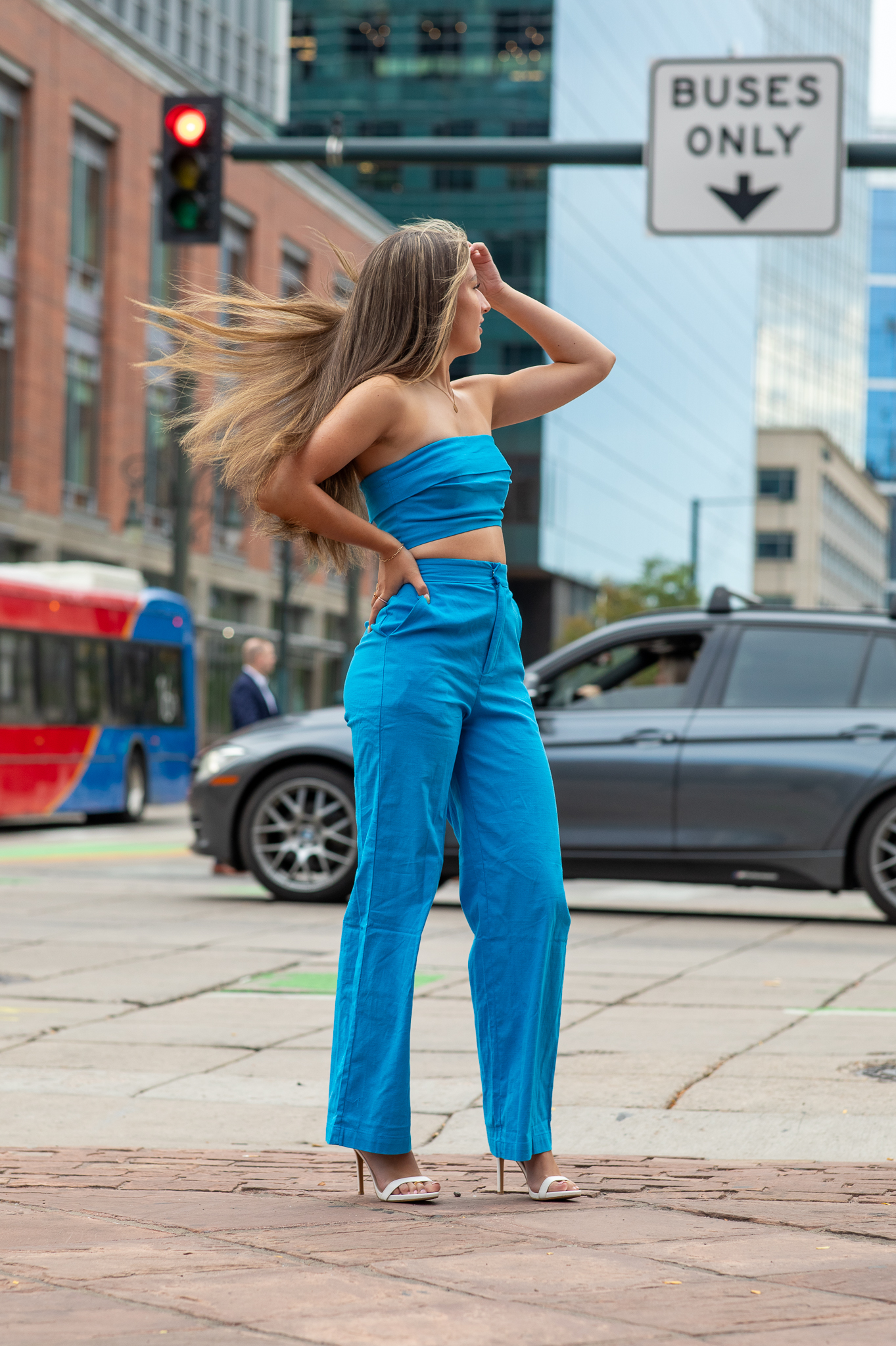 Young woman in a blue tube top and pants with long hair blowing, posing on a city sidewalk near traffic.