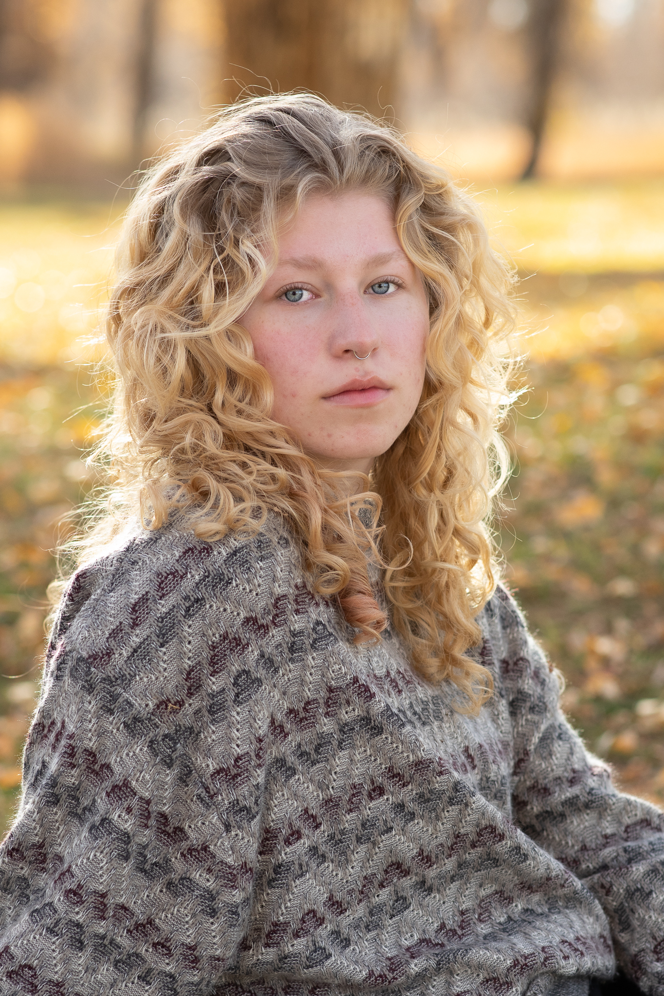Young woman with curly blonde hair wearing a patterned sweater, sitting outdoors on a sunny autumn day.