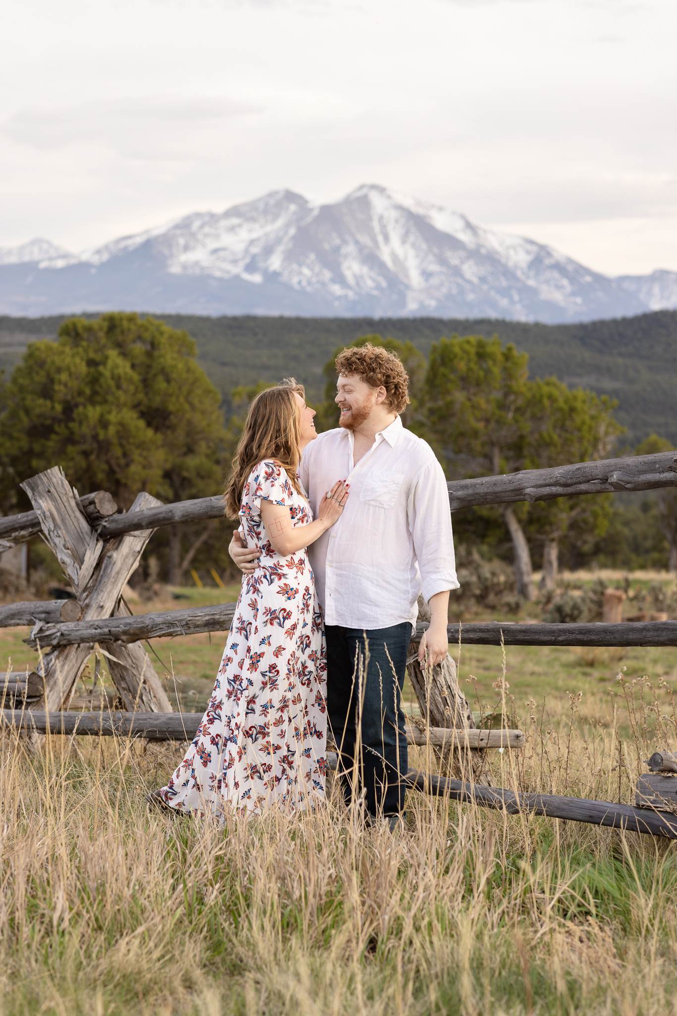 Couple embracing near a wooden fence with mountains and trees in the background.