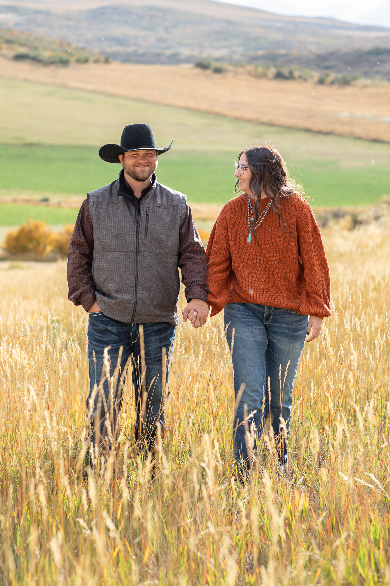 Couple holding hands and walking through a sunlit grassy field with rolling hills in the background.