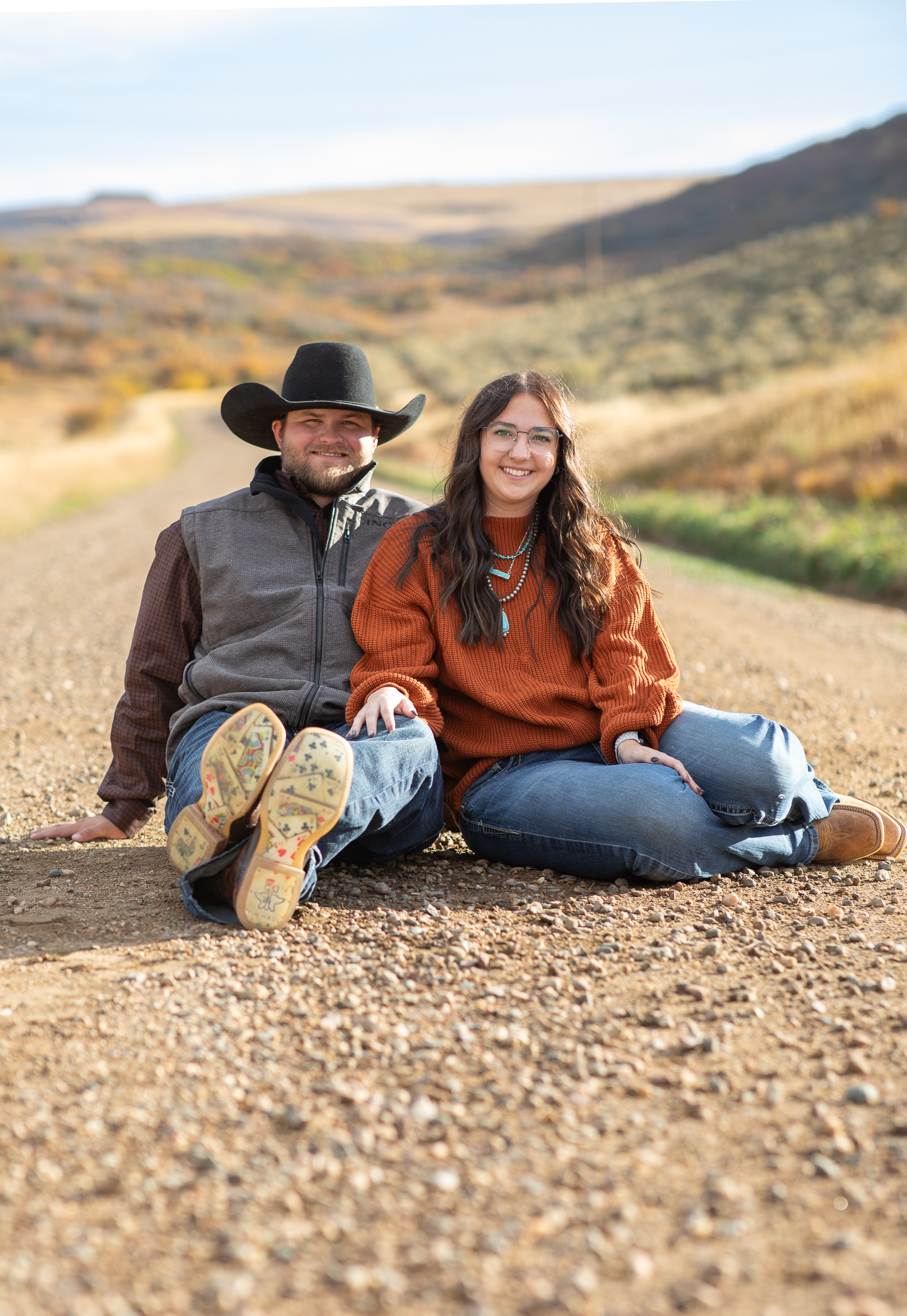 Smiling couple sitting side by side on a gravel road with scenic hills in the background.