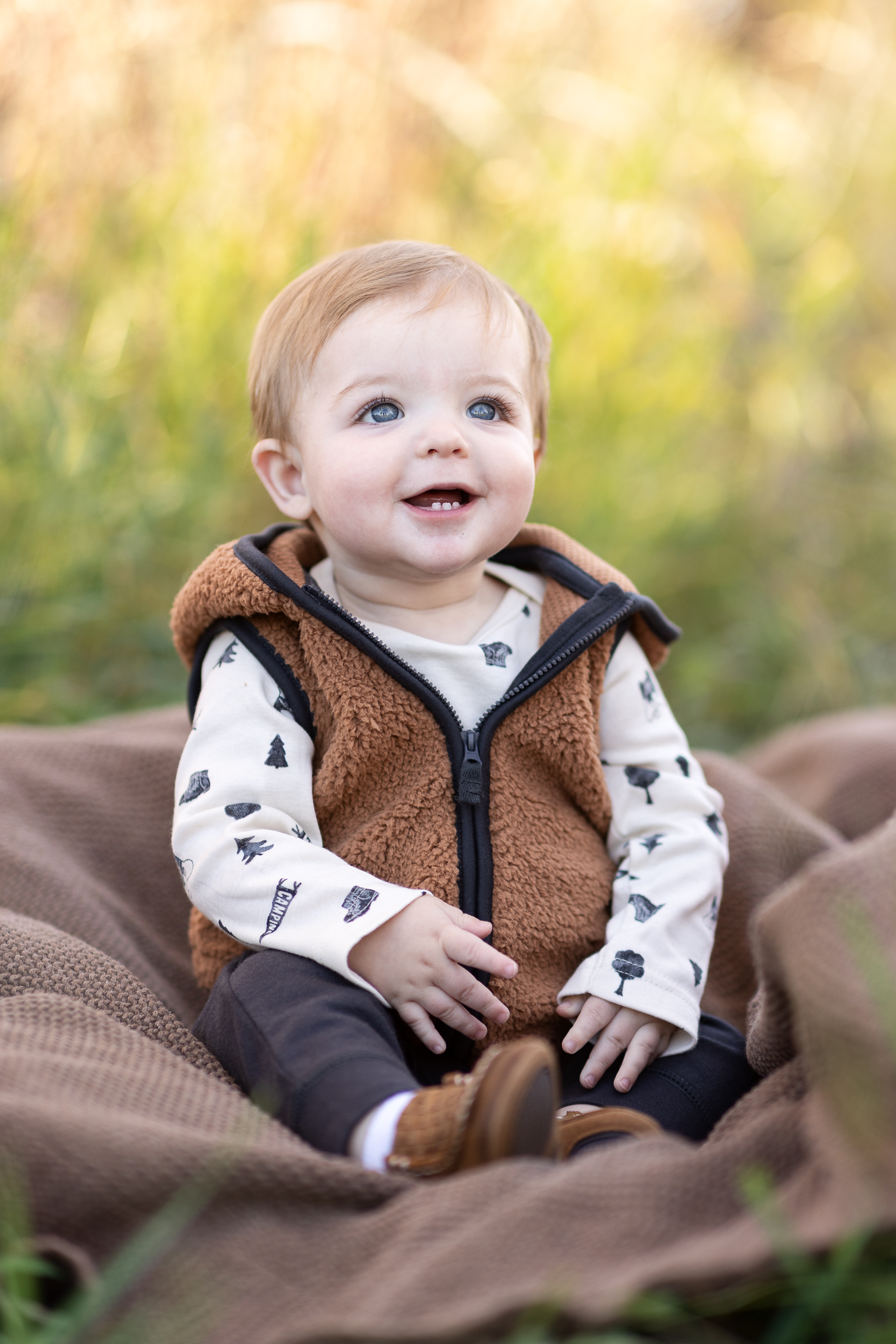 Smiling baby with blue eyes wearing a brown fleece vest sitting on a brown blanket outdoors.