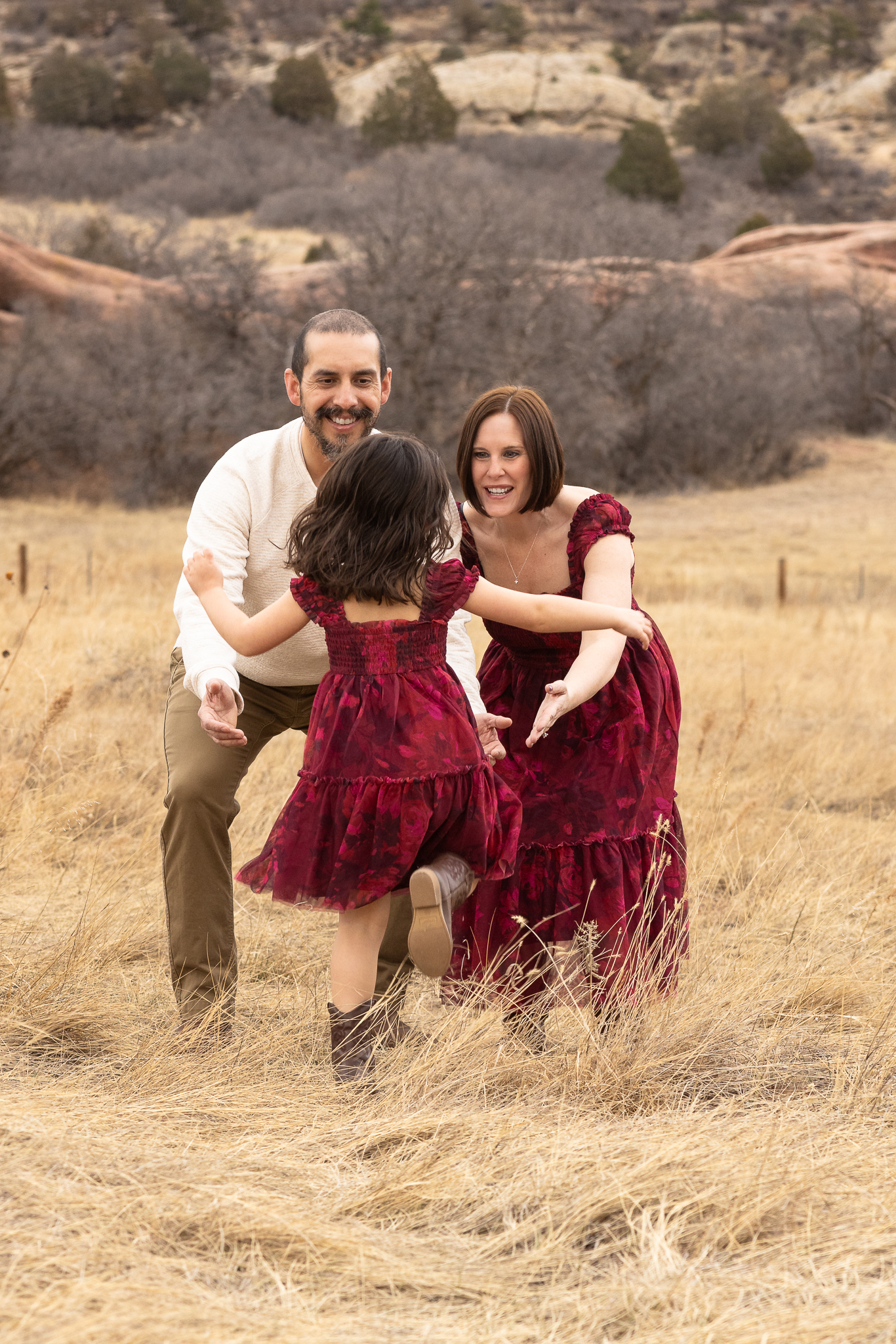 Young girl in a burgundy dress running toward her smiling parents with open arms in a dry grassy field.
