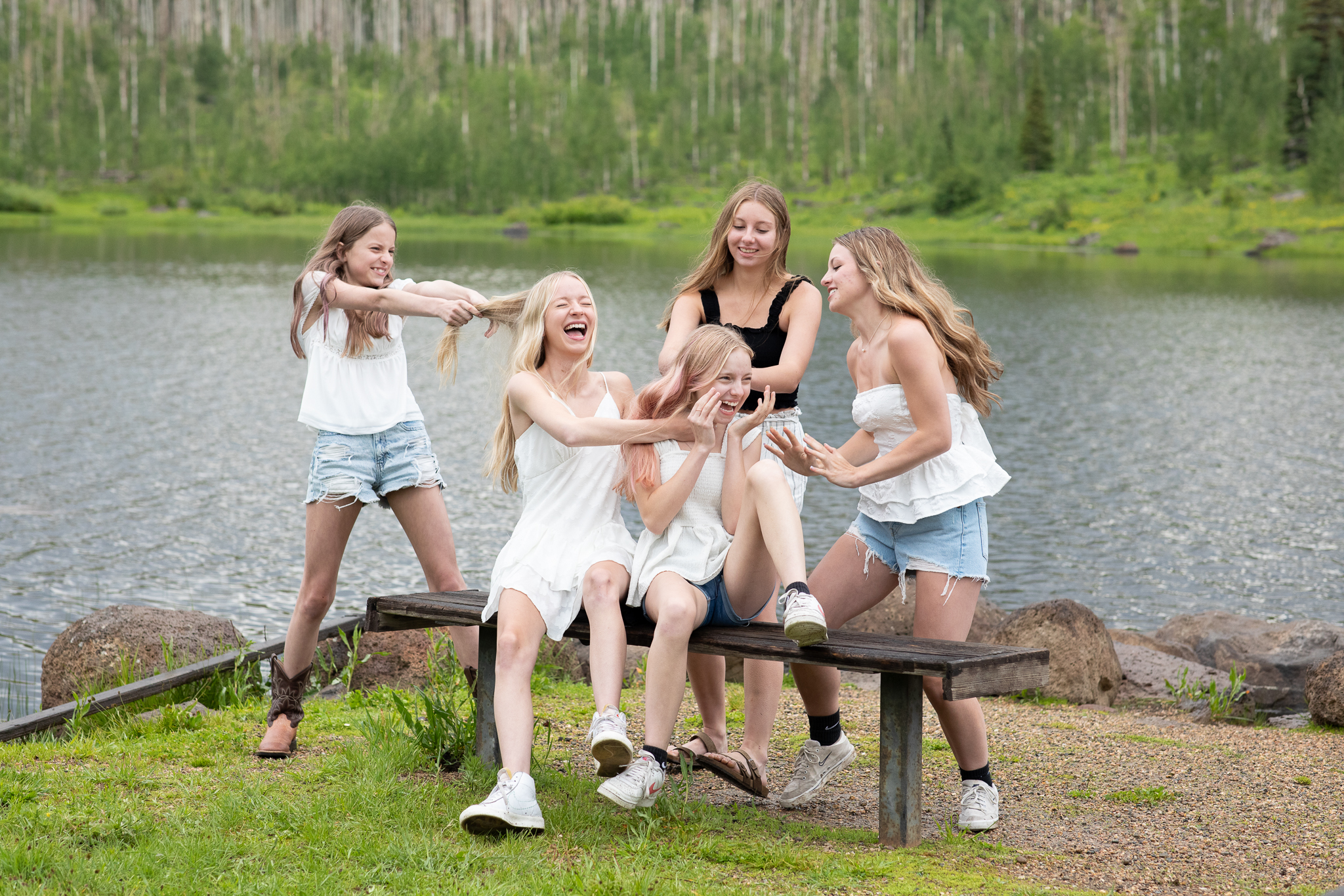 Five teenage girls laughing and playing on a bench by a lakeside with trees in the background.