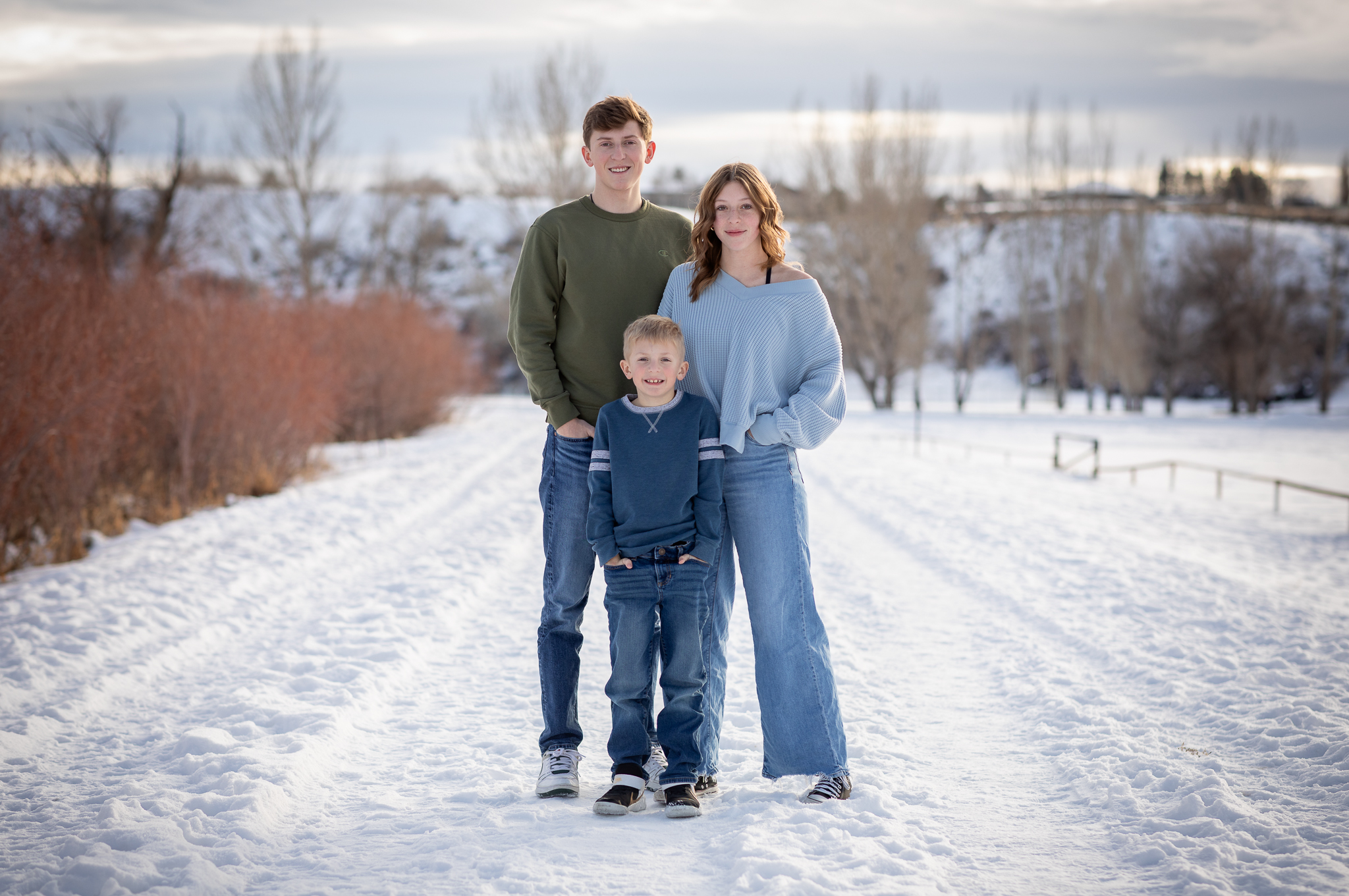 Three children standing on a snowy path outdoors in winter, smiling at the camera with bare trees and a cloudy sky in the background.