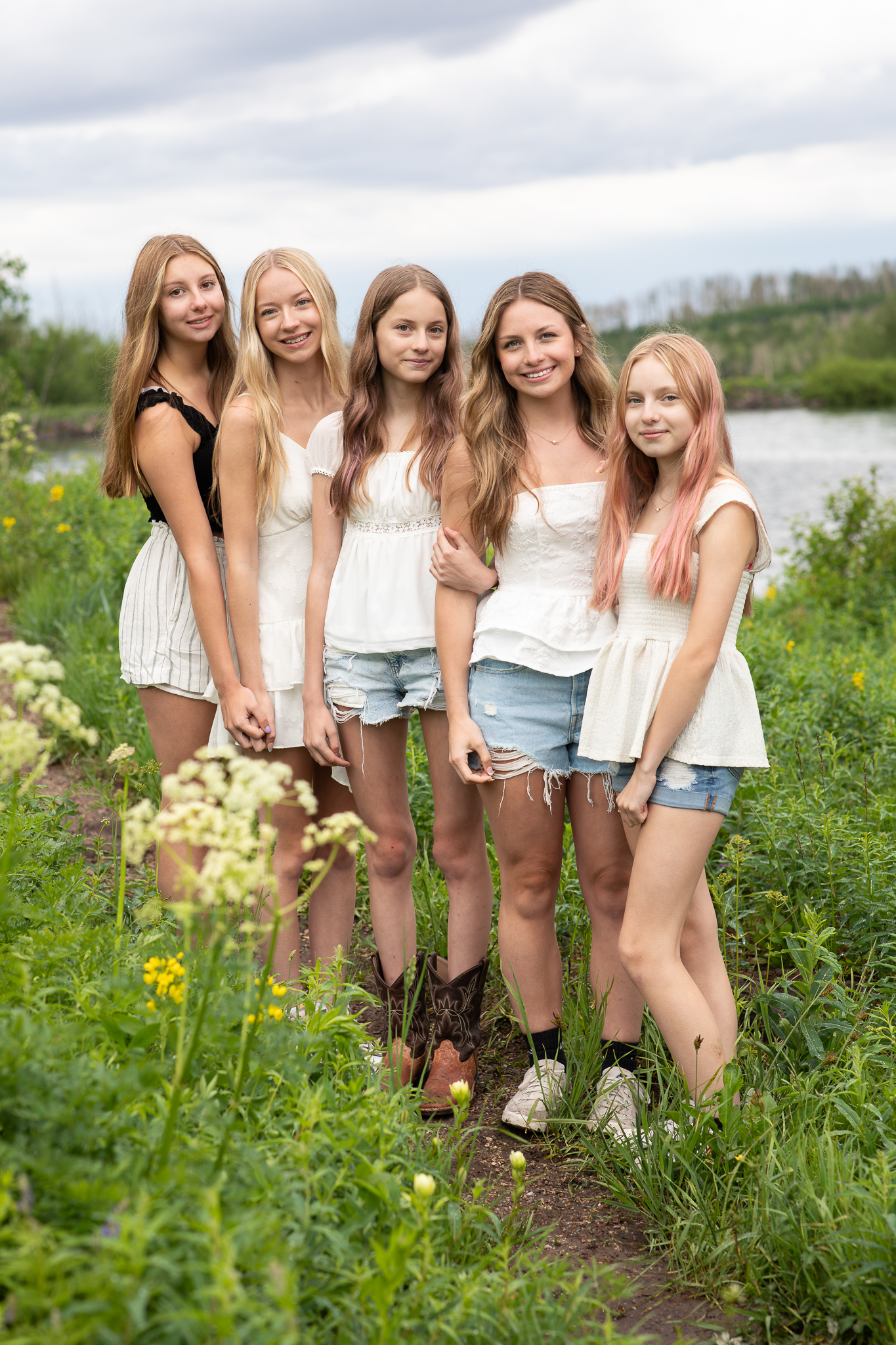 Five teenage girls standing close together on a wildflower-lined path near water, dressed in light summer outfits.