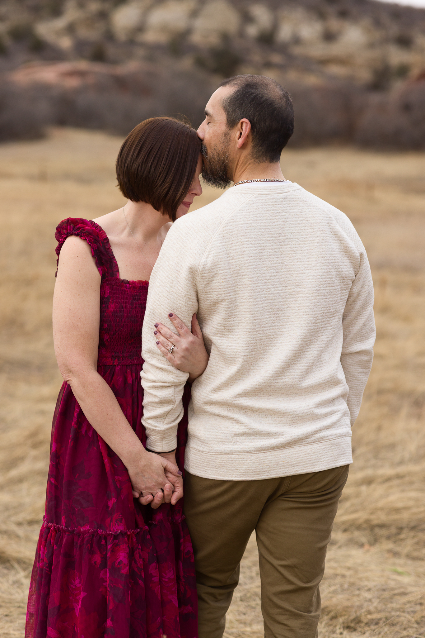 Man gently kissing a woman's forehead as they hold hands in a dry grassy field.