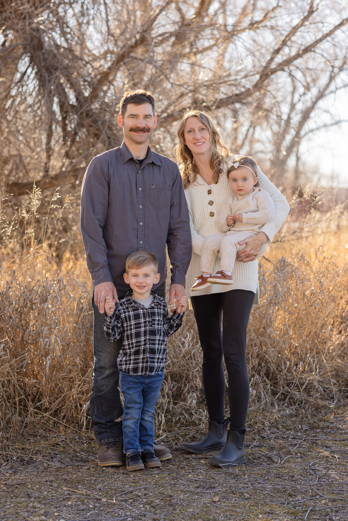 Family of four outdoors in autumn with father holding young boy’s hands and mother holding baby girl, standing in front of leafless trees and dry grass.