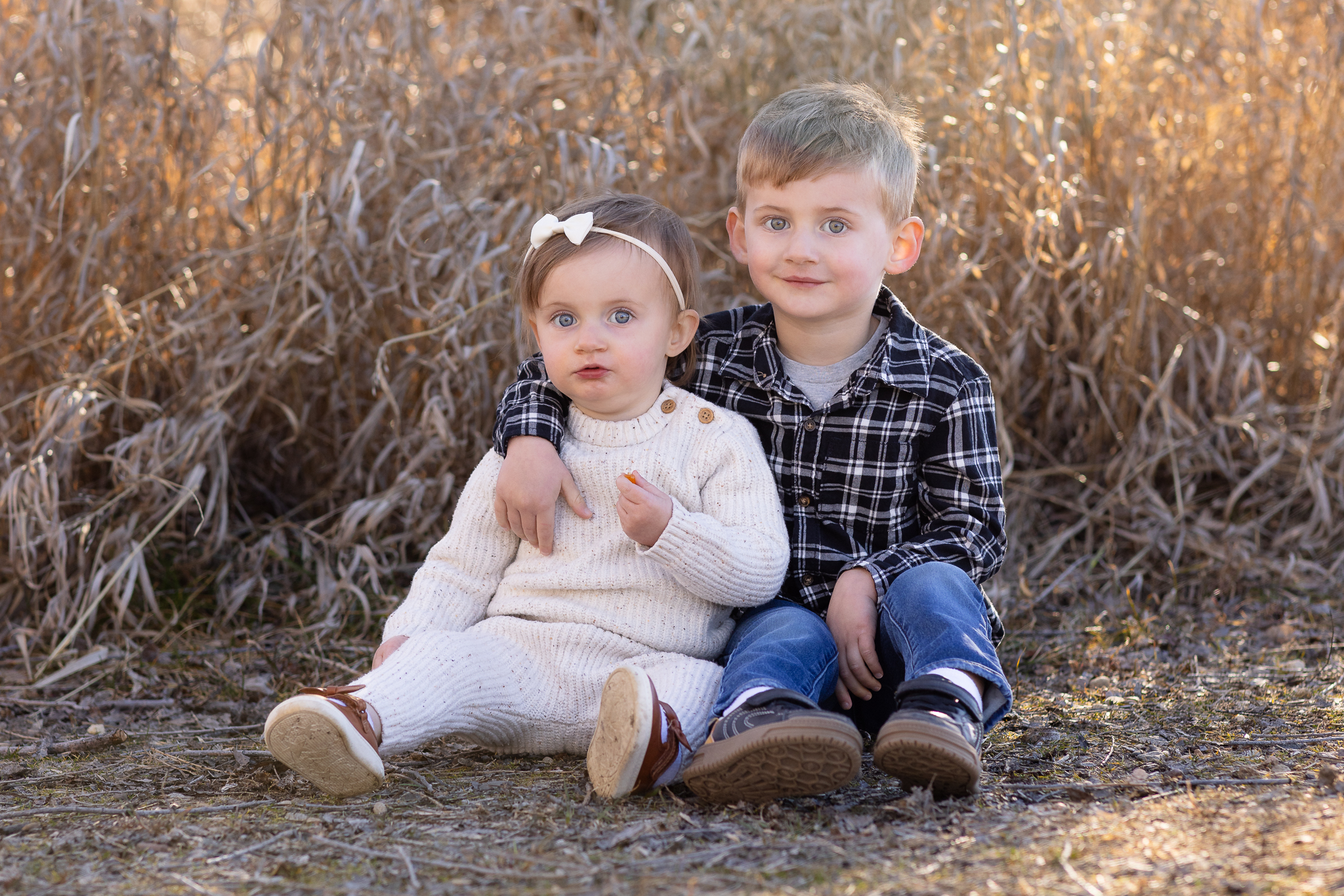 Young boy in a black plaid shirt with arm around a toddler girl in a white knit outfit sitting on the ground outdoors.