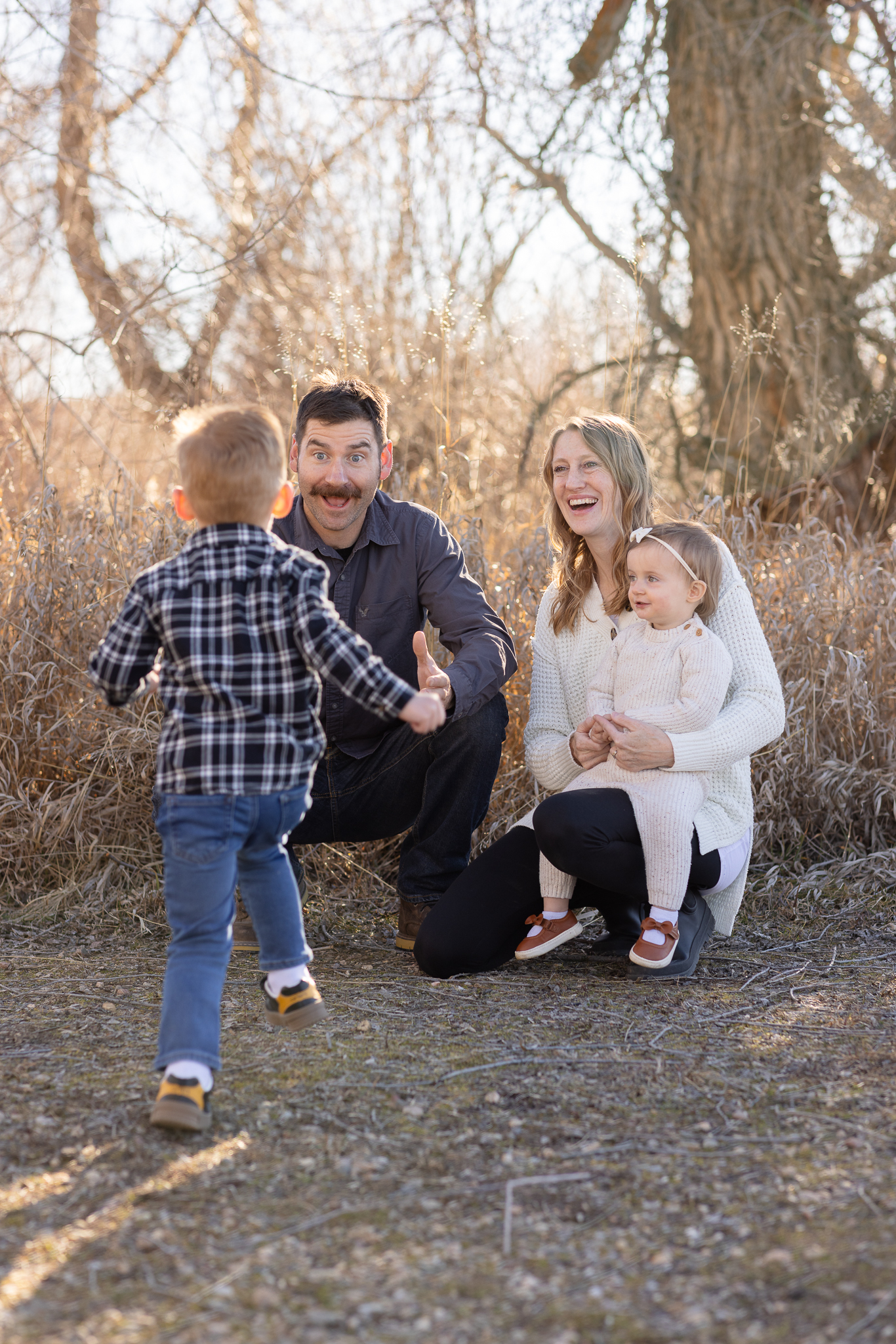 Young boy running towards a man with open arms while a woman holding a toddler smiles nearby outdoors.