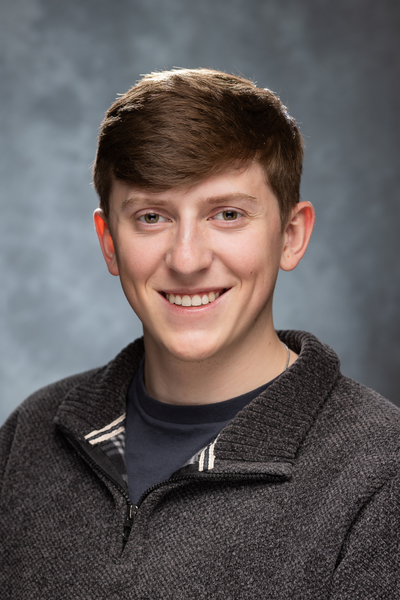 Smiling young man with brown hair wearing a dark gray sweater and black shirt against a gray background.
