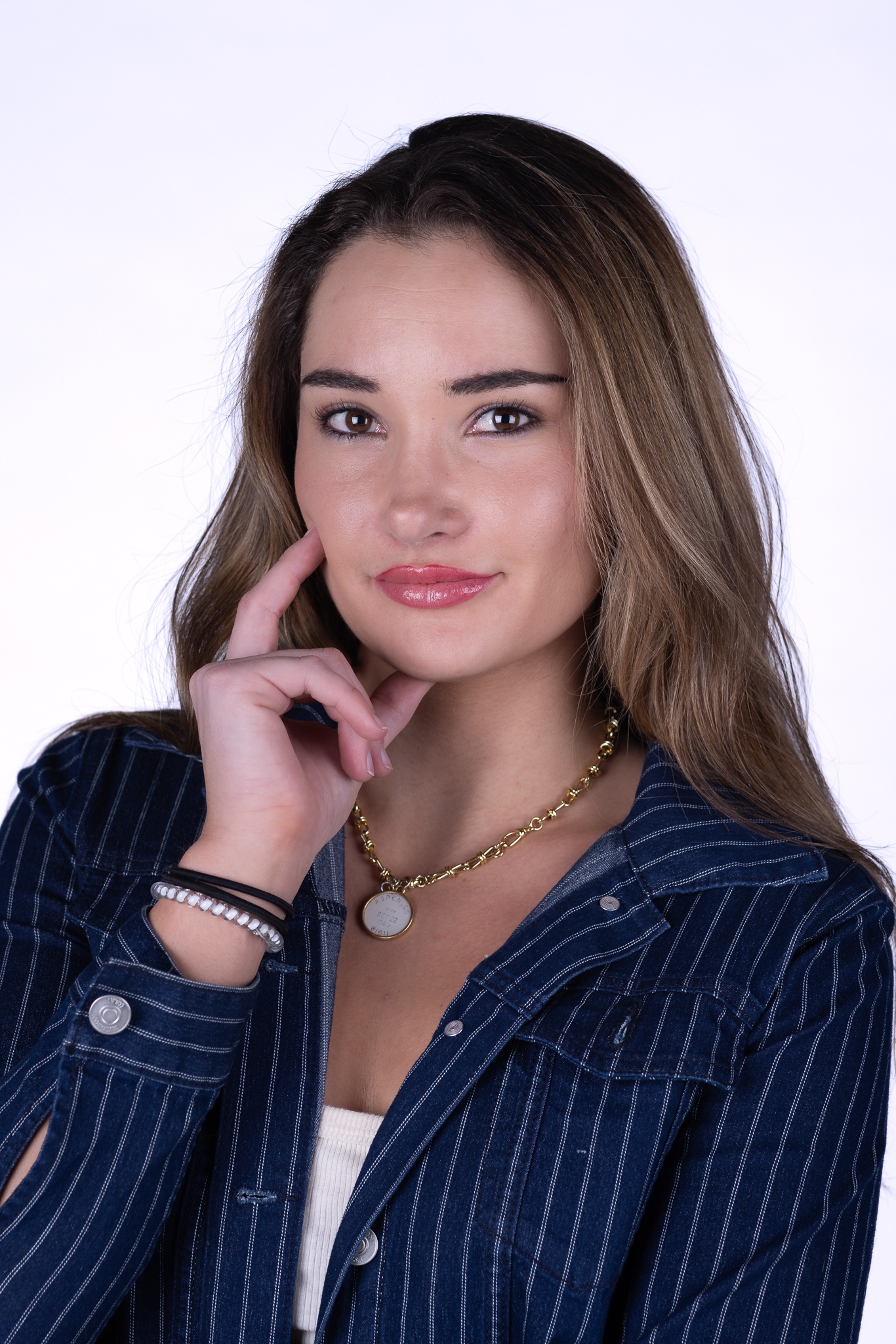 Portrait of a woman with long hair wearing a pinstriped denim jacket, a gold necklace, and thoughtful expression.