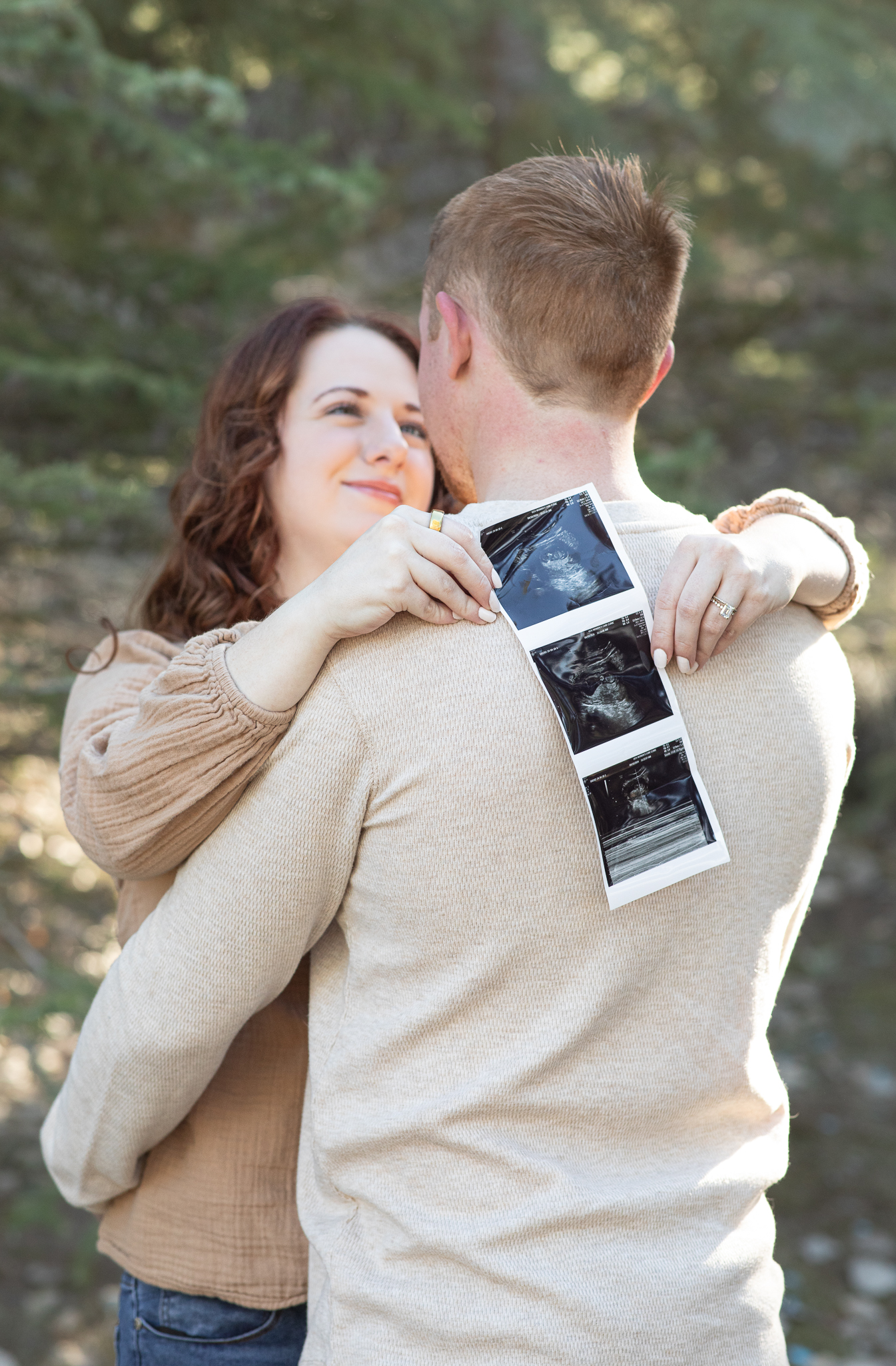Couple embracing outdoors with woman holding ultrasound images over man's shoulder.