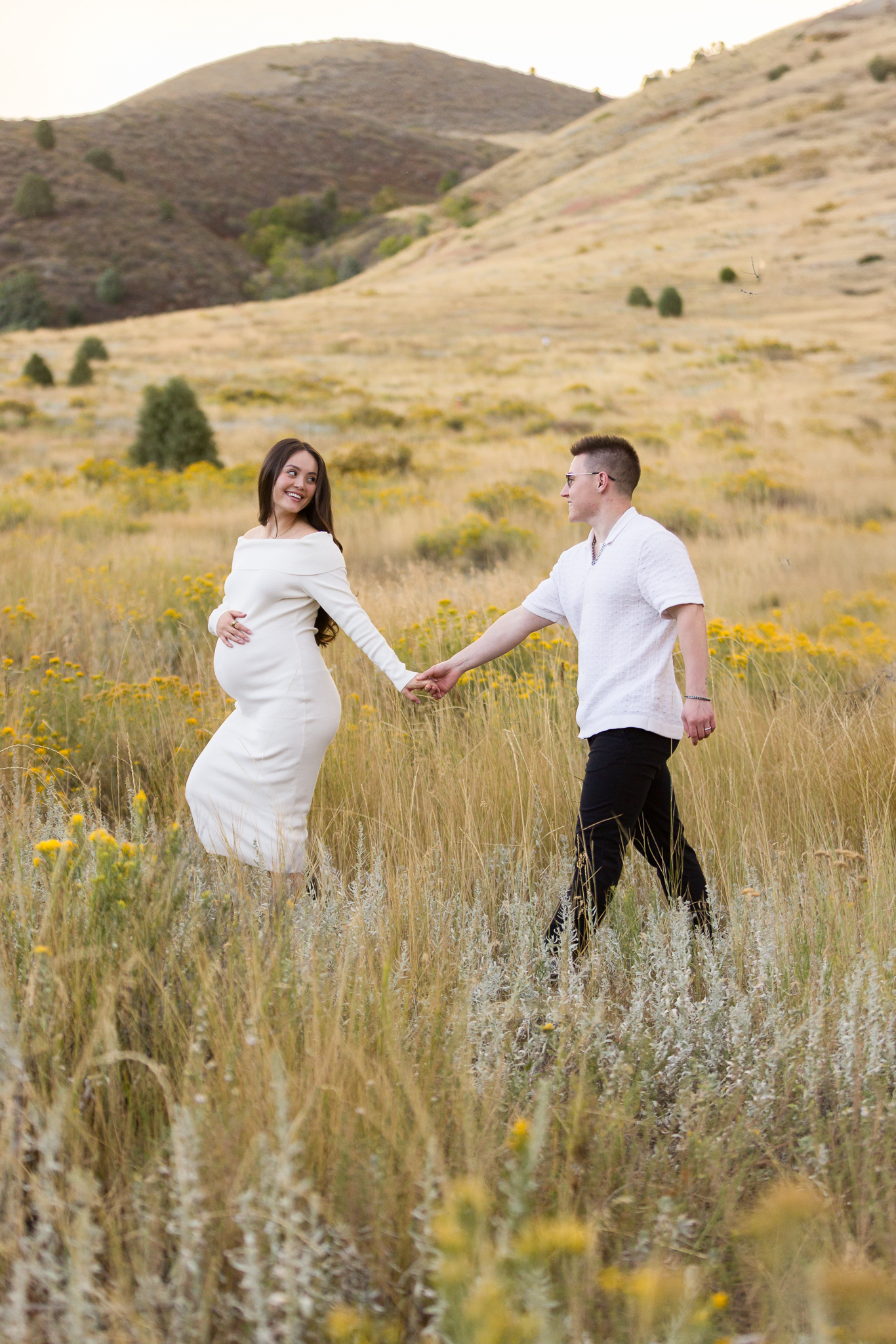 Pregnant woman in white dress holding hands with man in white shirt walking through a grassy field with yellow wildflowers and hills in the background.