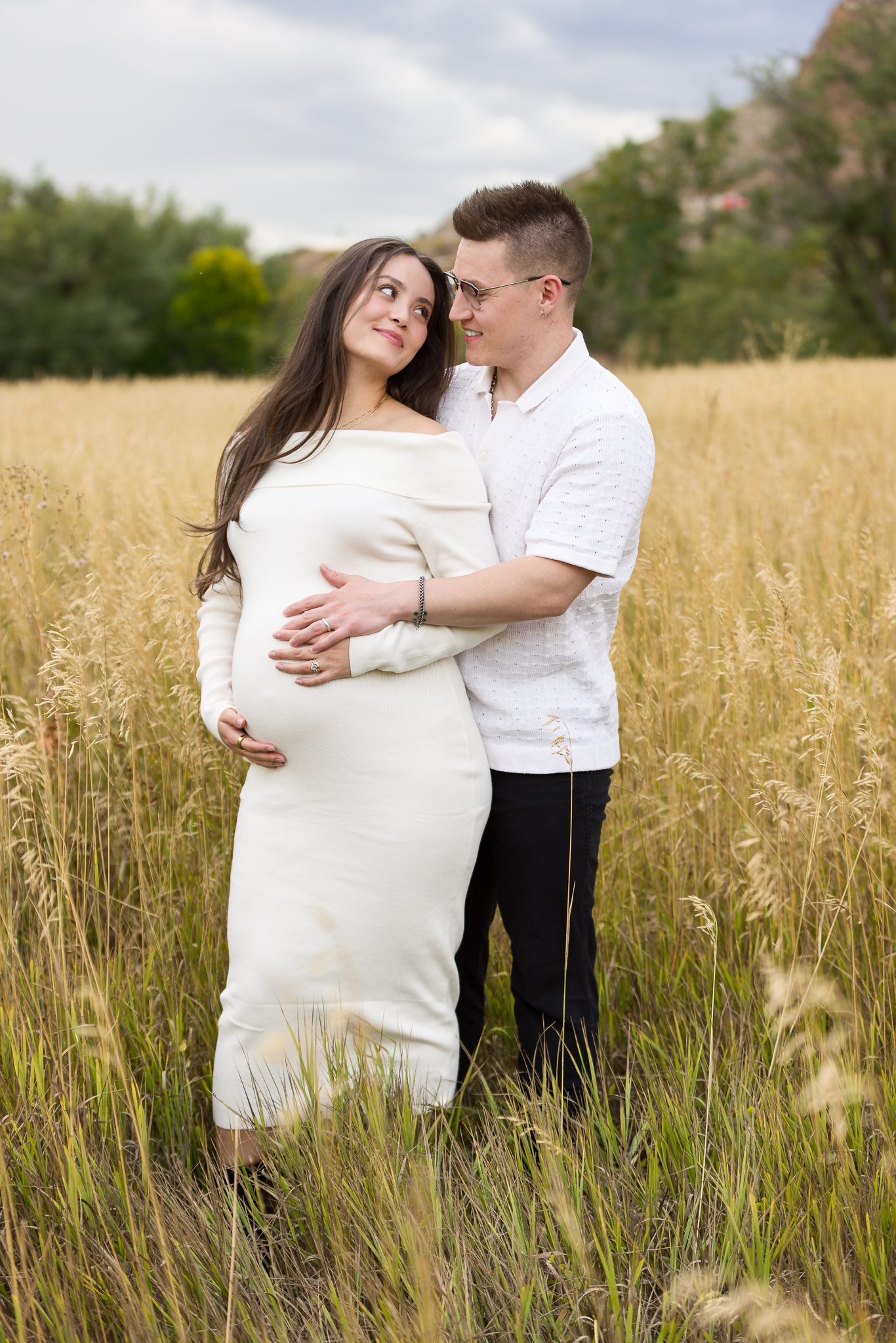 Pregnant woman in a white dress embraced by a man wearing glasses in a field of tall grass.