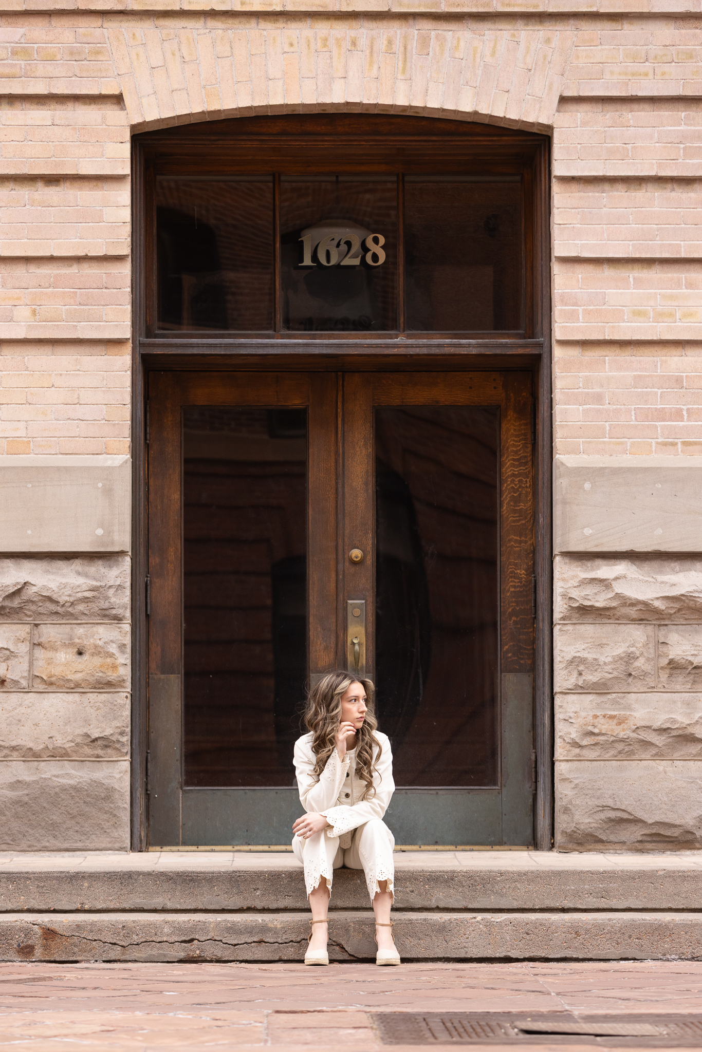 Young woman in white outfit sitting on stone steps in front of wooden double doors with the number 1628 above.