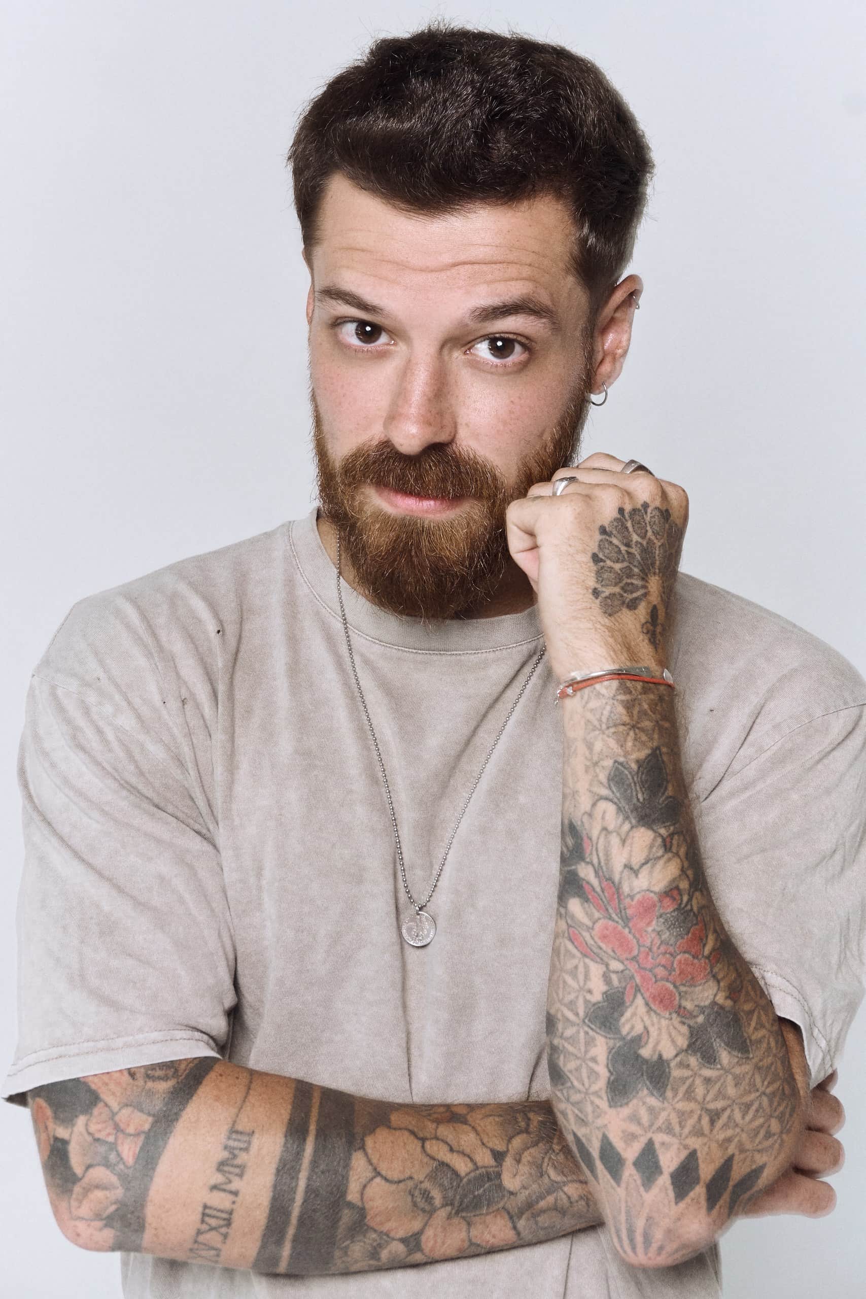 Man with medium-length wavy hair, round glasses, and a beard wearing a brown cardigan and white shirt against a dark background.
