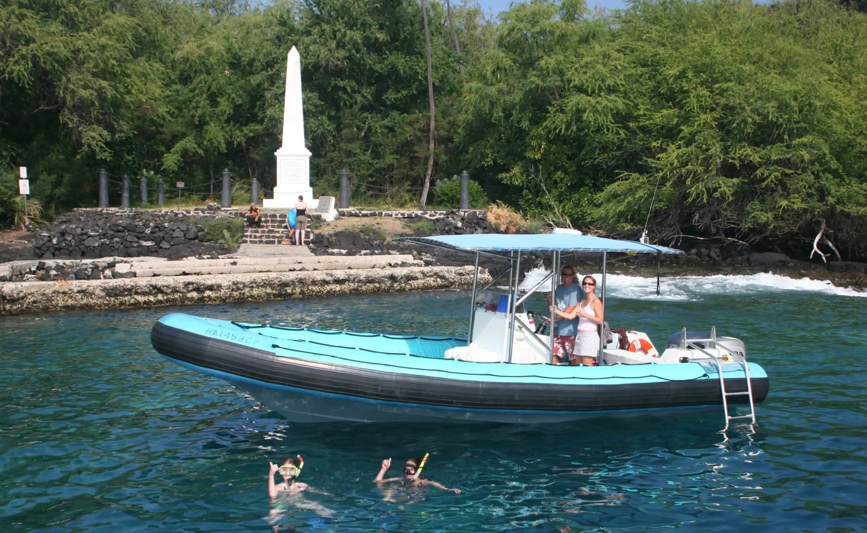 Two snorkelers in the water near a blue inflatable boat with two people onboard by a shoreline monument.