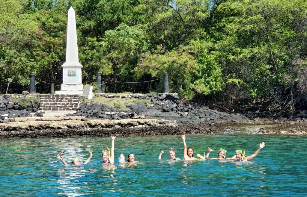 Seven people waving while snorkeling in clear blue water near a stone monument and green trees on shore.