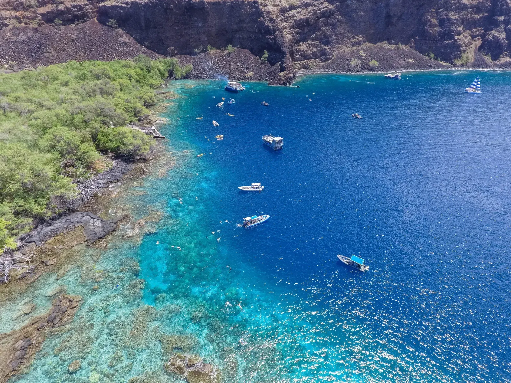 Aerial view of clear blue ocean with boats near a rocky shoreline with green vegetation.
