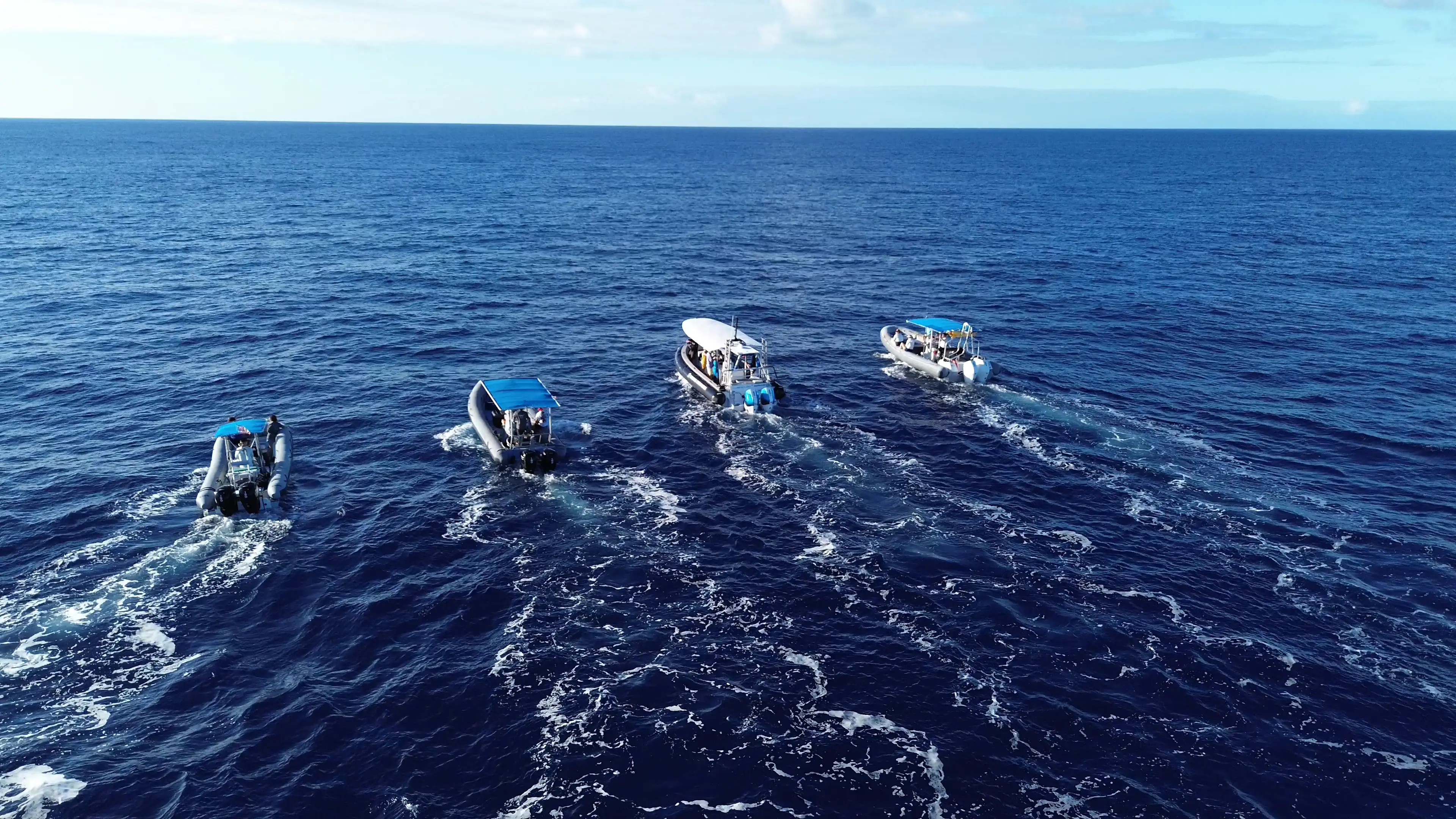 Four small motorboats with blue tops moving forward on a calm blue ocean under a partly cloudy sky.