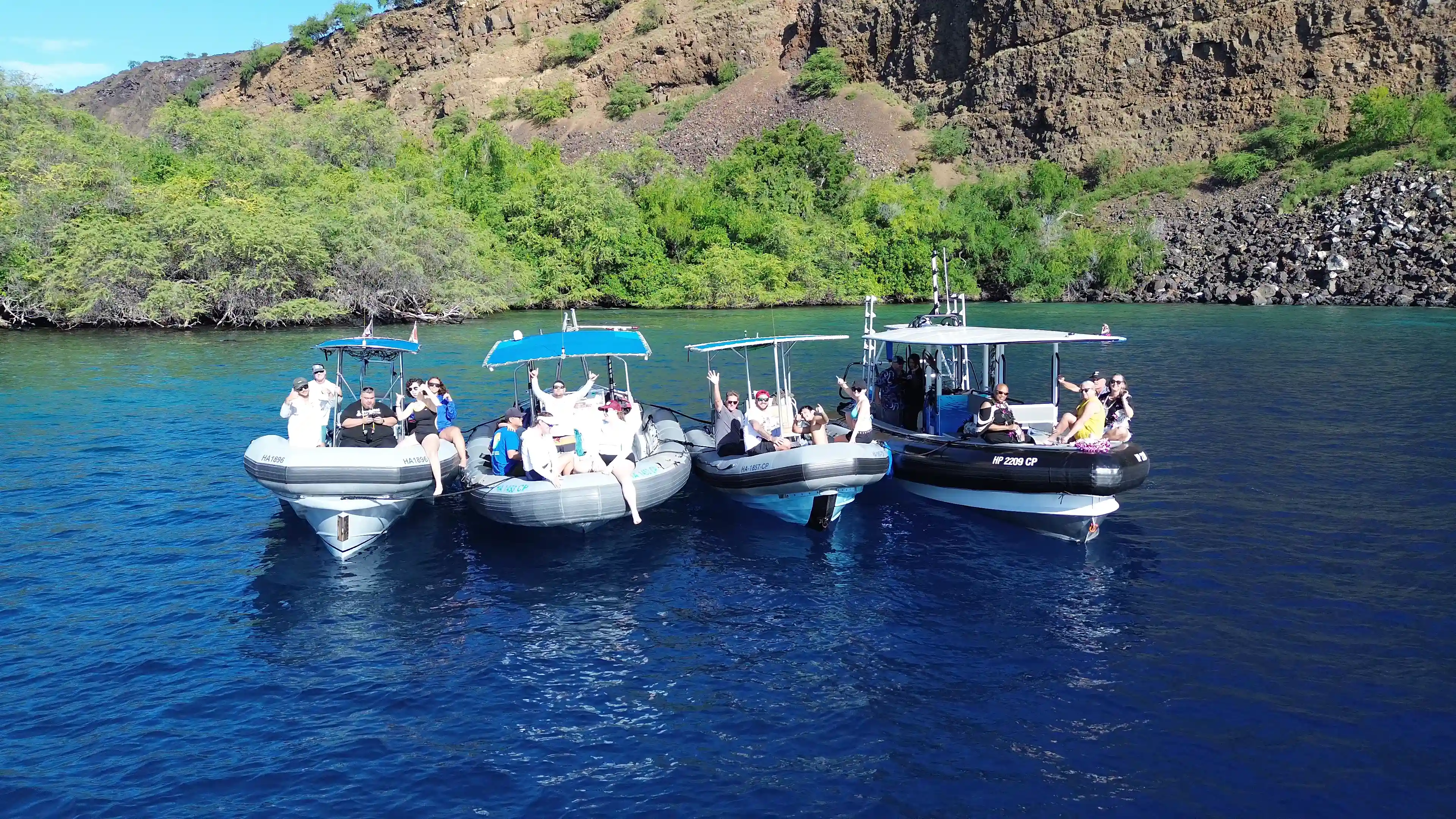 Four small boats with people waving and relaxing on blue water near rocky shore with green trees.