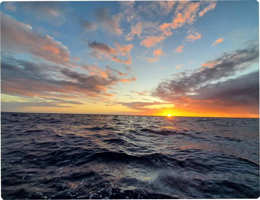 Sun setting over a calm ocean with scattered clouds illuminated in orange and pink.