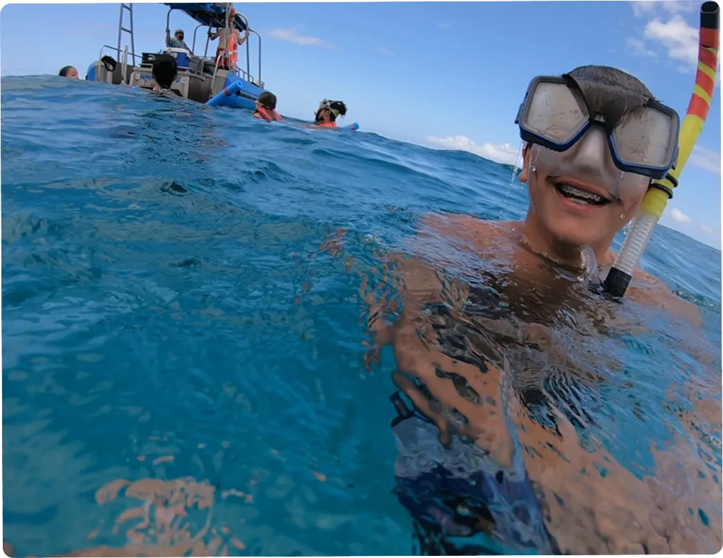 Smiling person with braces snorkeling in clear blue ocean water near a boat with other snorkelers.