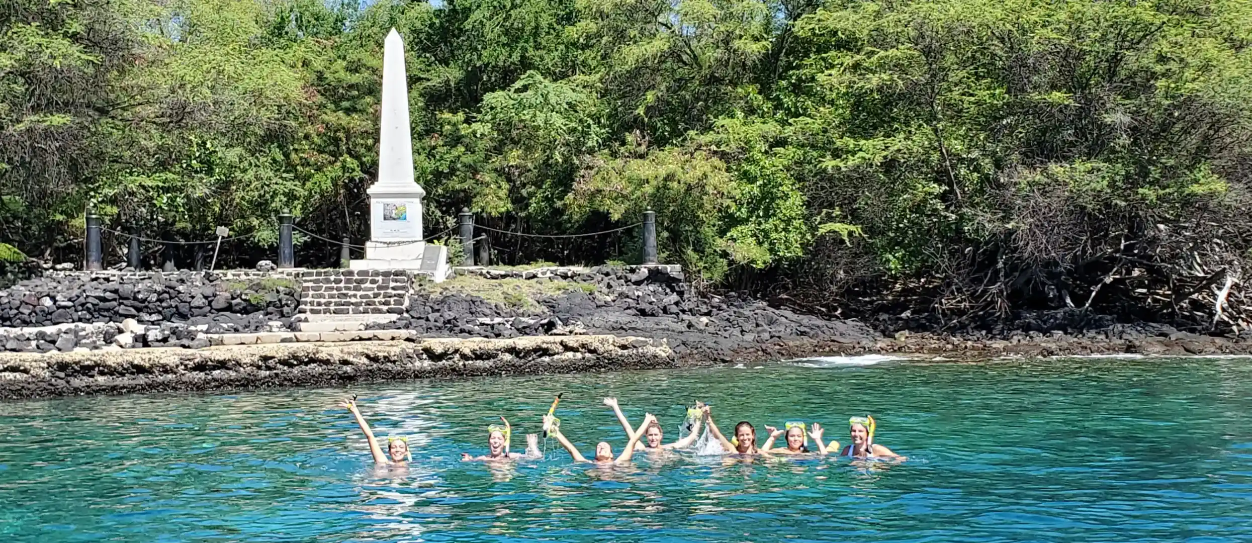 Group of snorkelers waving in clear blue water near rocky shore and tall white monument surrounded by trees.