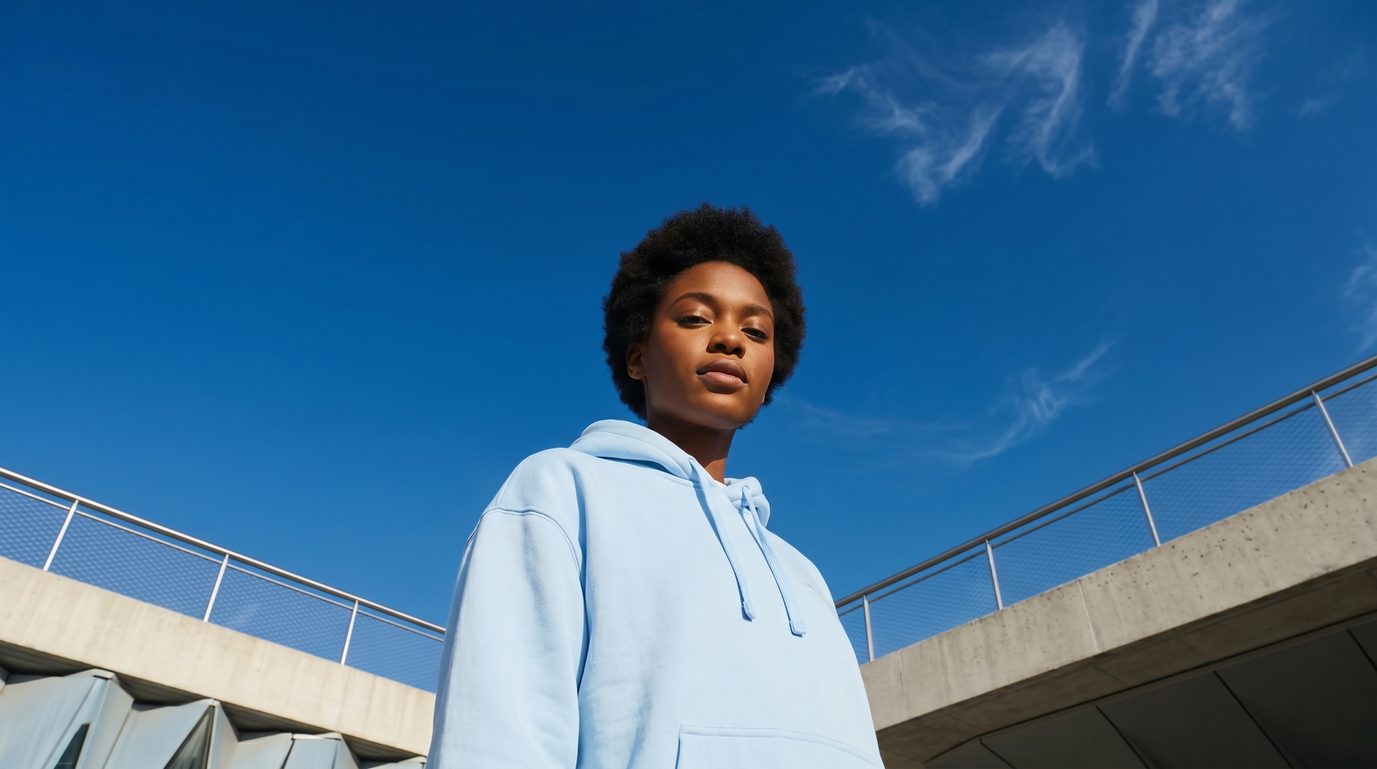 Confident young woman in a light blue hoodie standing outdoors under a clear blue sky with concrete railing in the background.