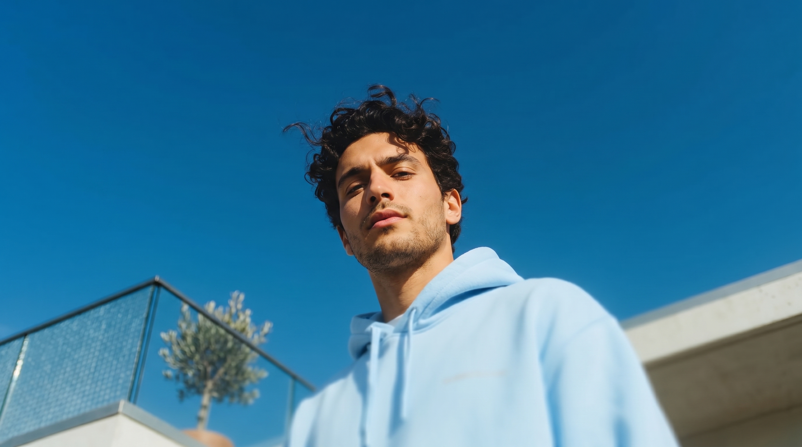 Young man with curly hair wearing a light blue hoodie standing outdoors against a clear blue sky.