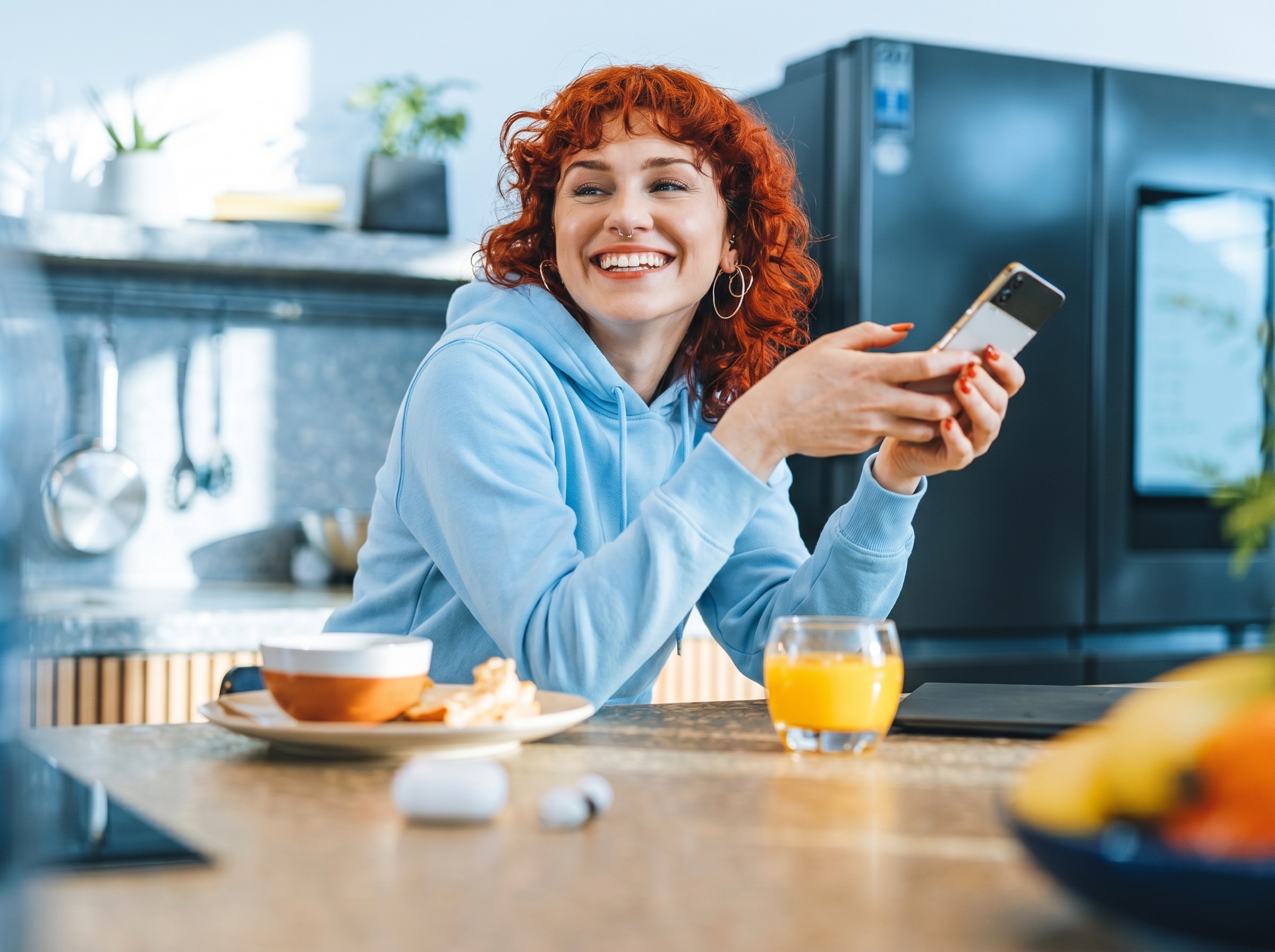 A smiling young woman with curly red hair in a blue hoodie holding a smartphone while sitting at a kitchen table with a glass of orange juice and a bowl of food.