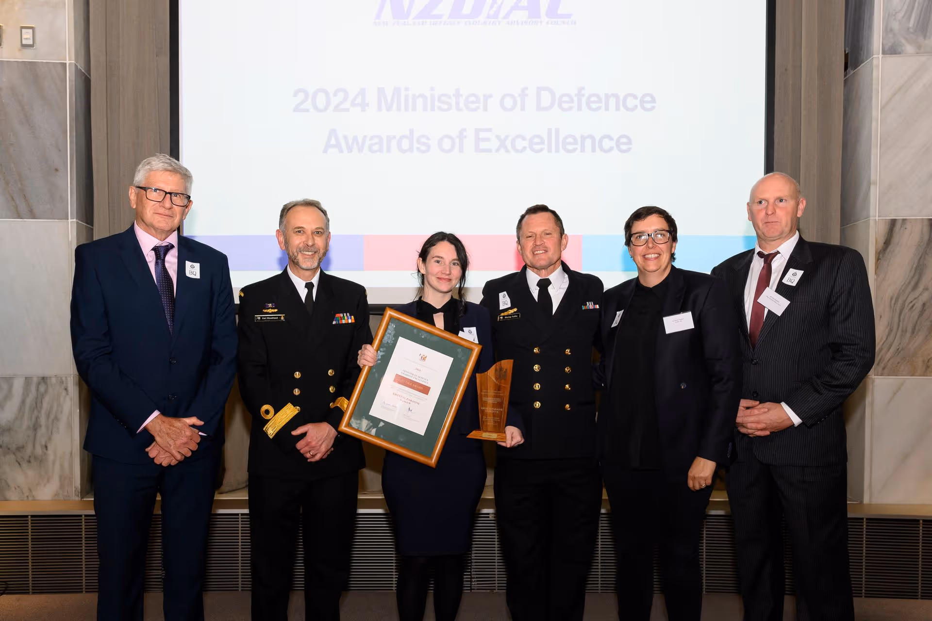 Six people standing in a row at the 2024 Minister of Defence Awards of Excellence, with one woman holding a framed certificate and a wooden trophy.