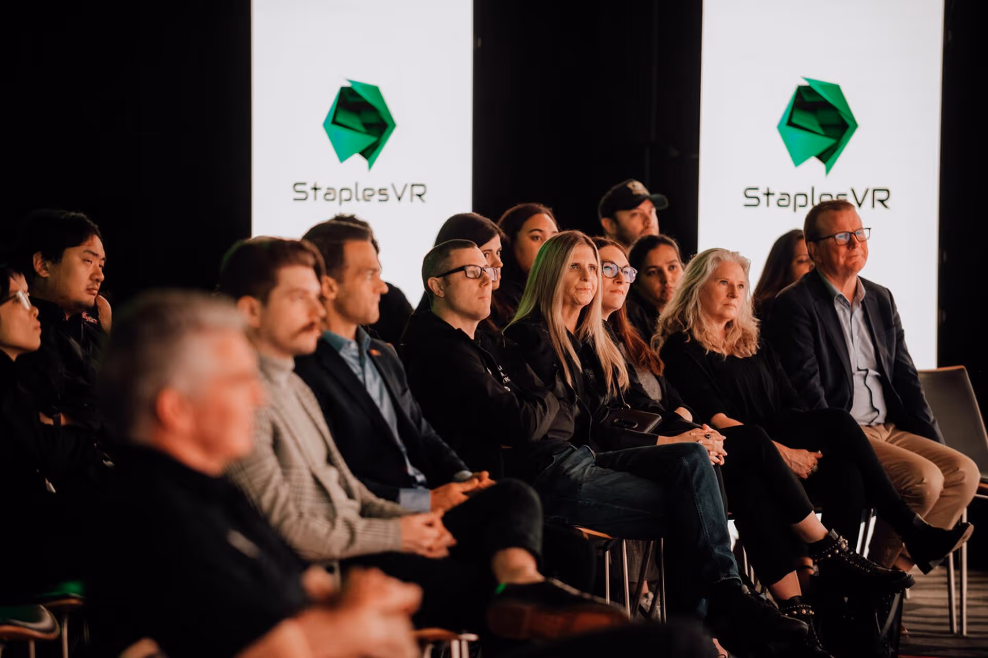 Audience seated in a dimly lit room attentively watching an event with two large screens displaying the StaplesVR logo in the background.